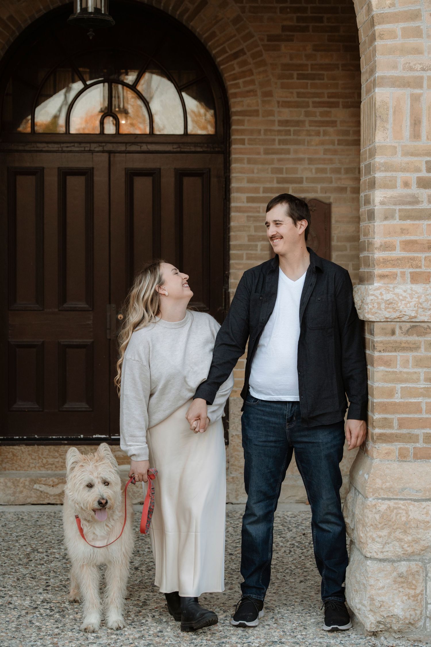 A smiling couple holds hands with a white dog on a red leash, standing before an arched wooden door.