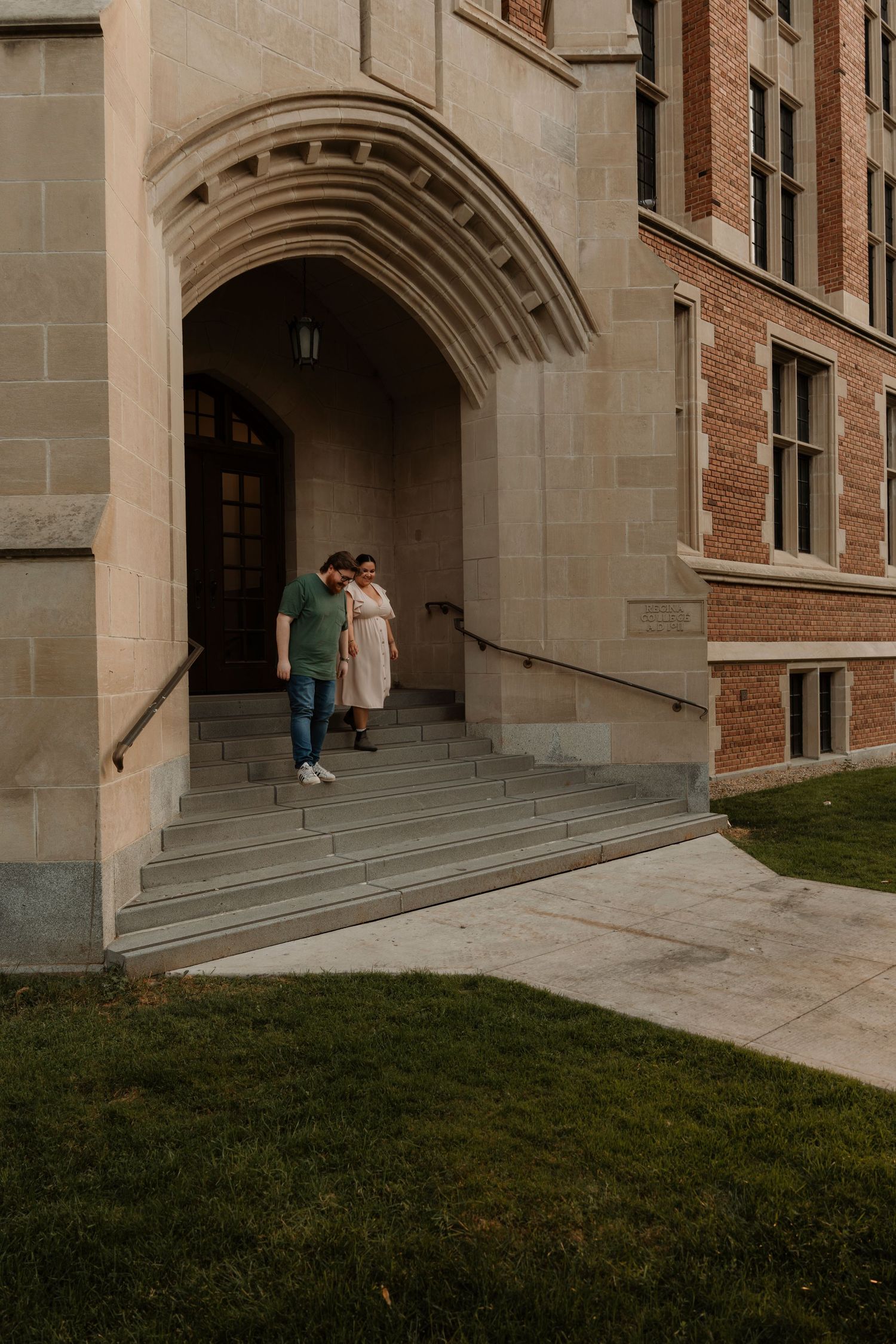 Couple walks down stone steps under Gothic arch entrance of historic brick university building.