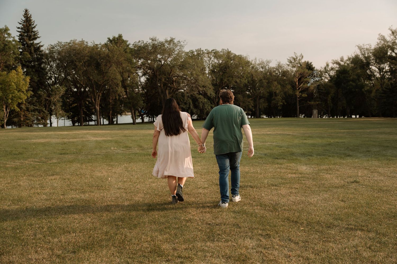 A couple walks hand-in-hand across a lush green park, surrounded by tall trees under a cloudy sky.