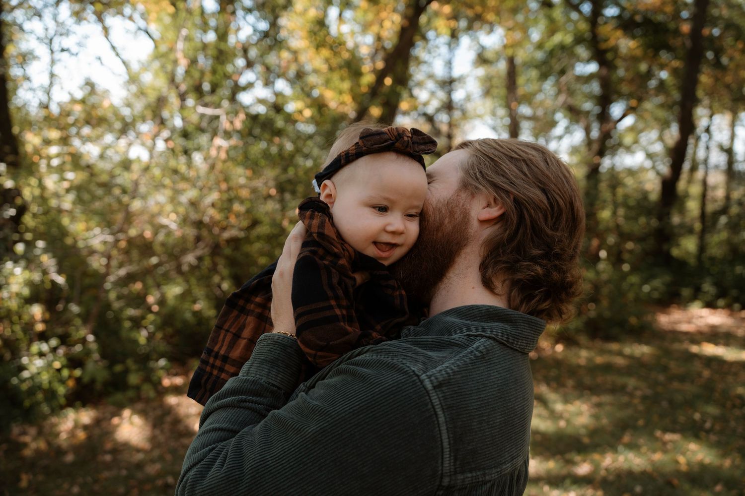 A father tenderly kisses his smiling baby in a sunlit autumn forest setting.