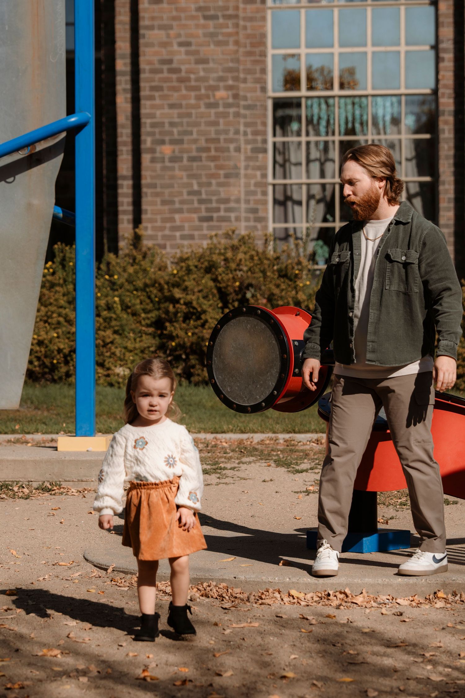 A bearded father walks with his young daughter in an orange skirt at a sunny playground near a brick building.