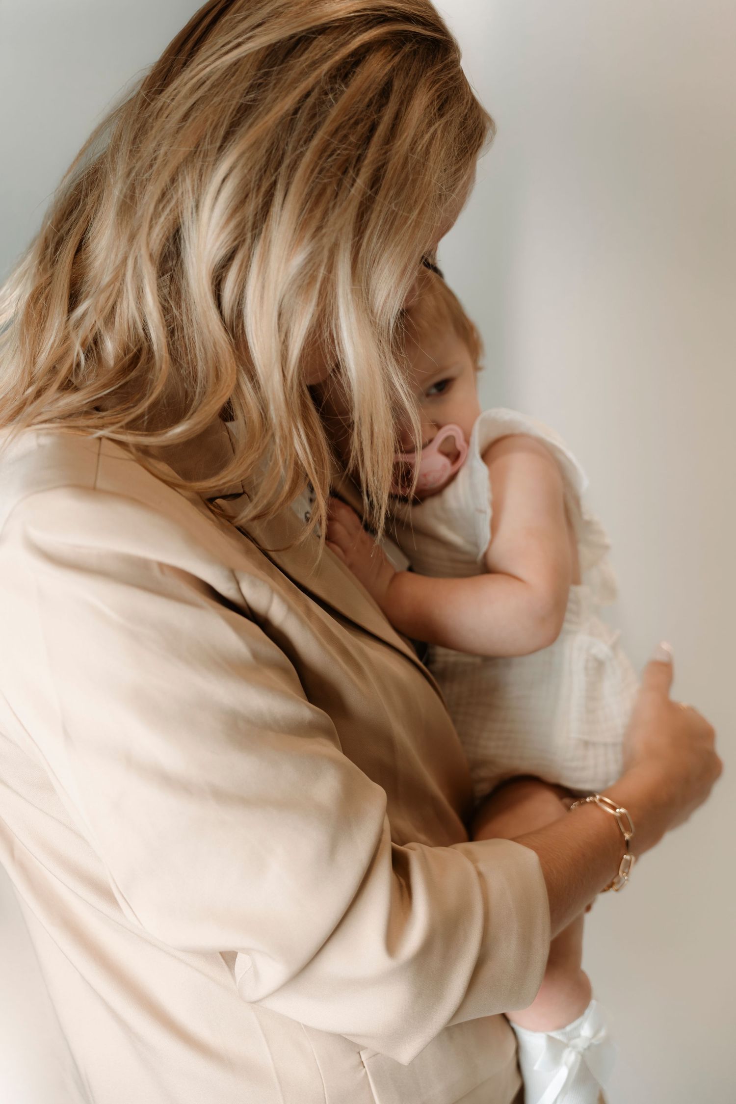 Blonde woman in beige blazer tenderly holding a baby dressed in white, sharing a gentle embrace.
