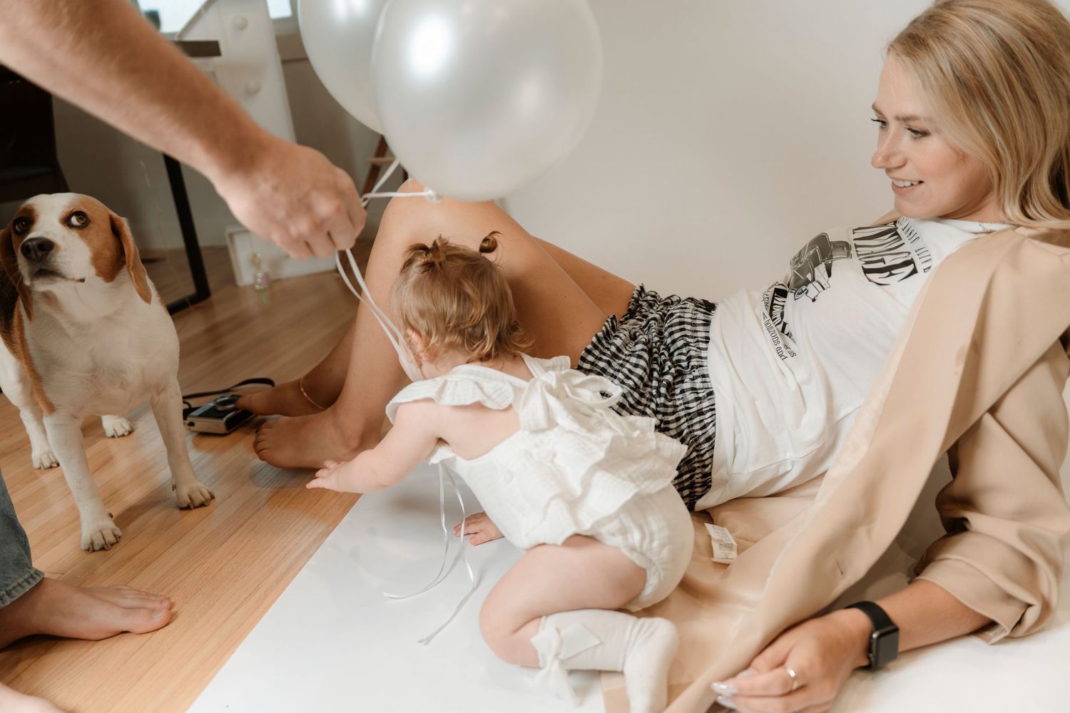 A woman sits on the floor with a baby and a beagle dog, holding white balloons in a bright room.