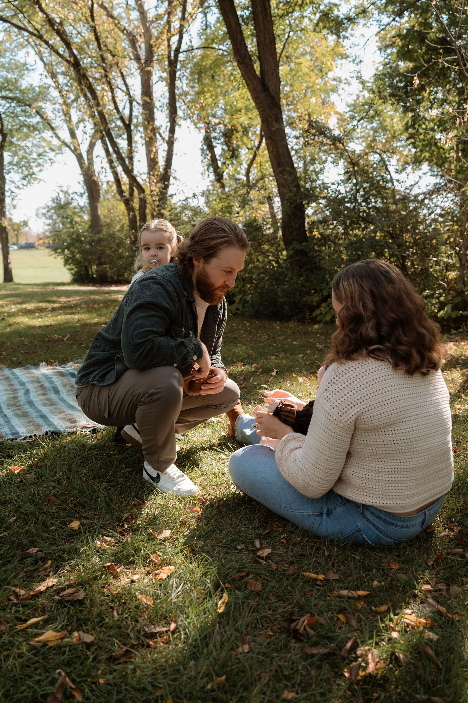 Family enjoying a picnic in a sunny park, sitting on grass surrounded by autumn trees and fallen leaves.