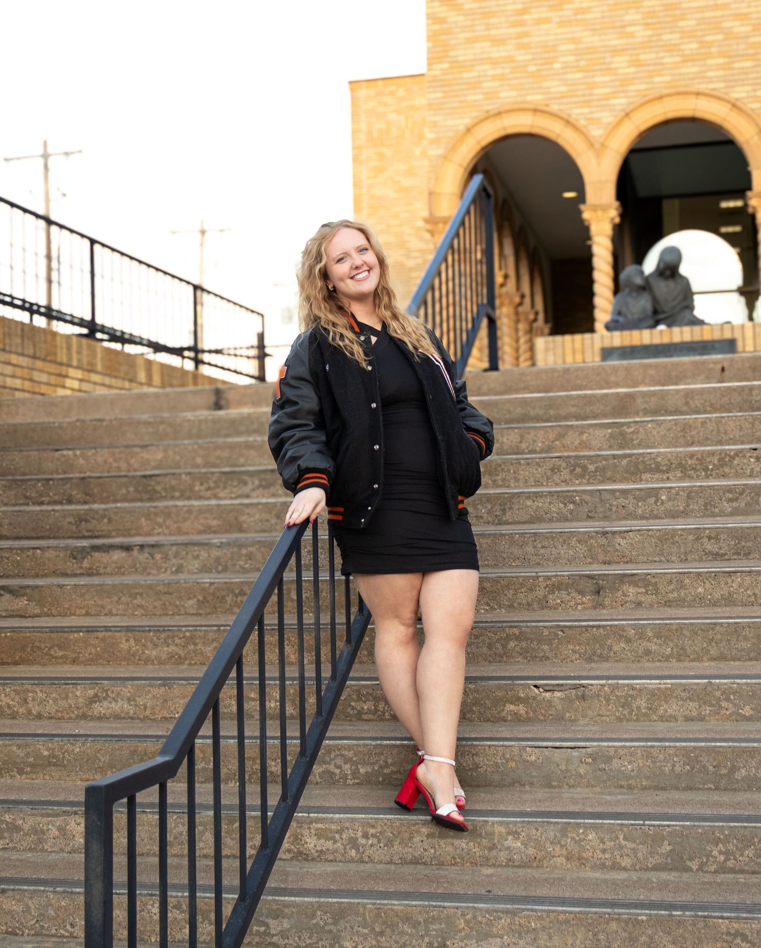 Young woman in black mini dress and varsity jacket poses on outdoor steps wearing red heels, smiling confidently.Senior Photos inspo