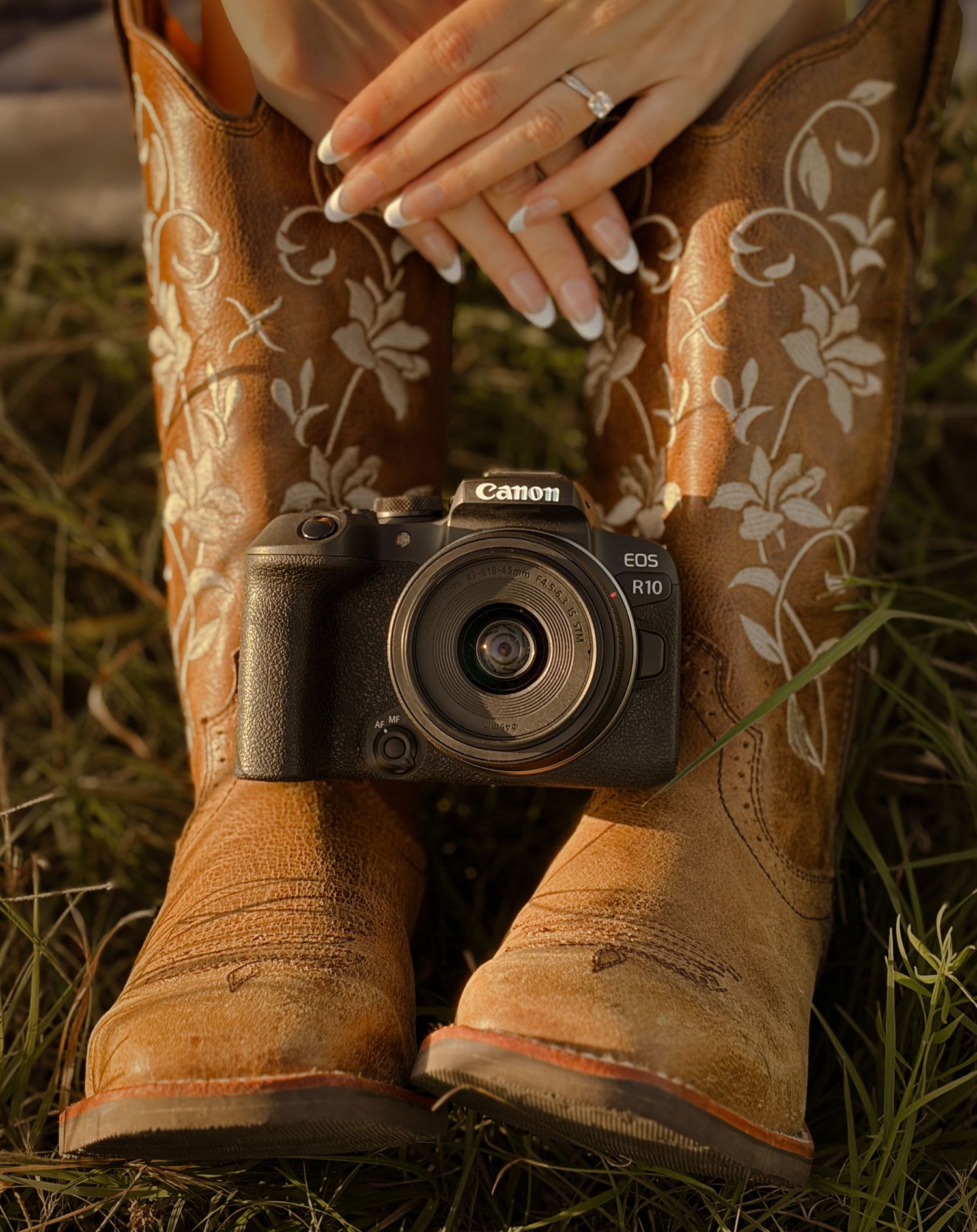 Canon EOS R10 camera resting between embroidered tan cowboy boots with manicured hands visible above. Branding inspo