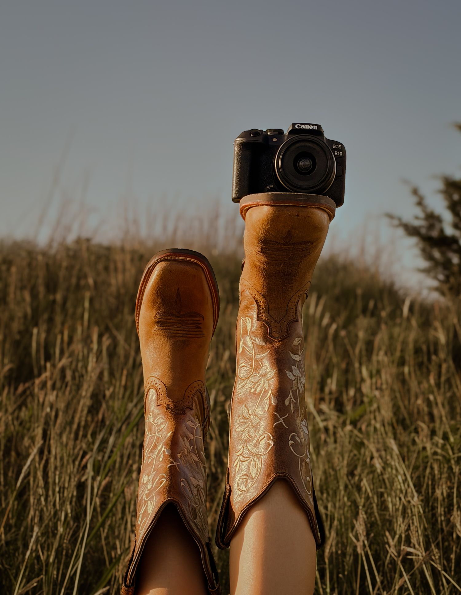 lying in grass field with legs raised wearing tan cowboy boots, balancing a Canon camera on top. Branding inspo
