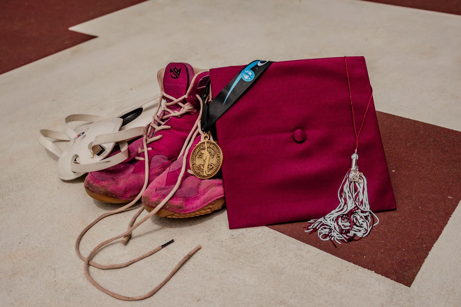 Maroon graduation cap, medal, and pink wrestling shoes arranged on a floor for a cap and gown session.Senior Photos inspo