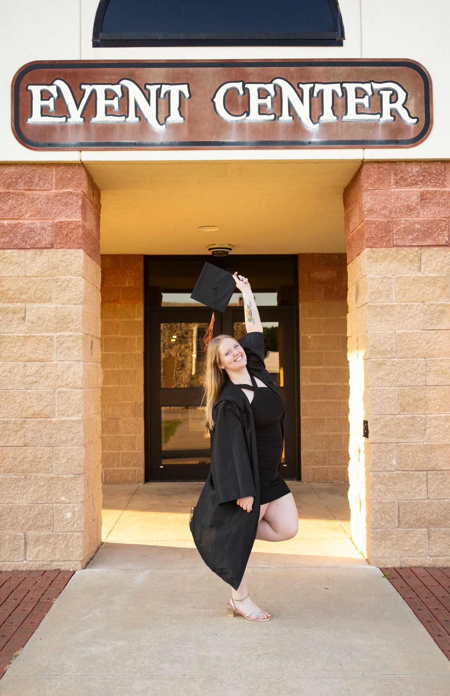 A graduate in black cap and gown joyfully tosses her cap outside an Event Center entrance. Senior Photos inspo