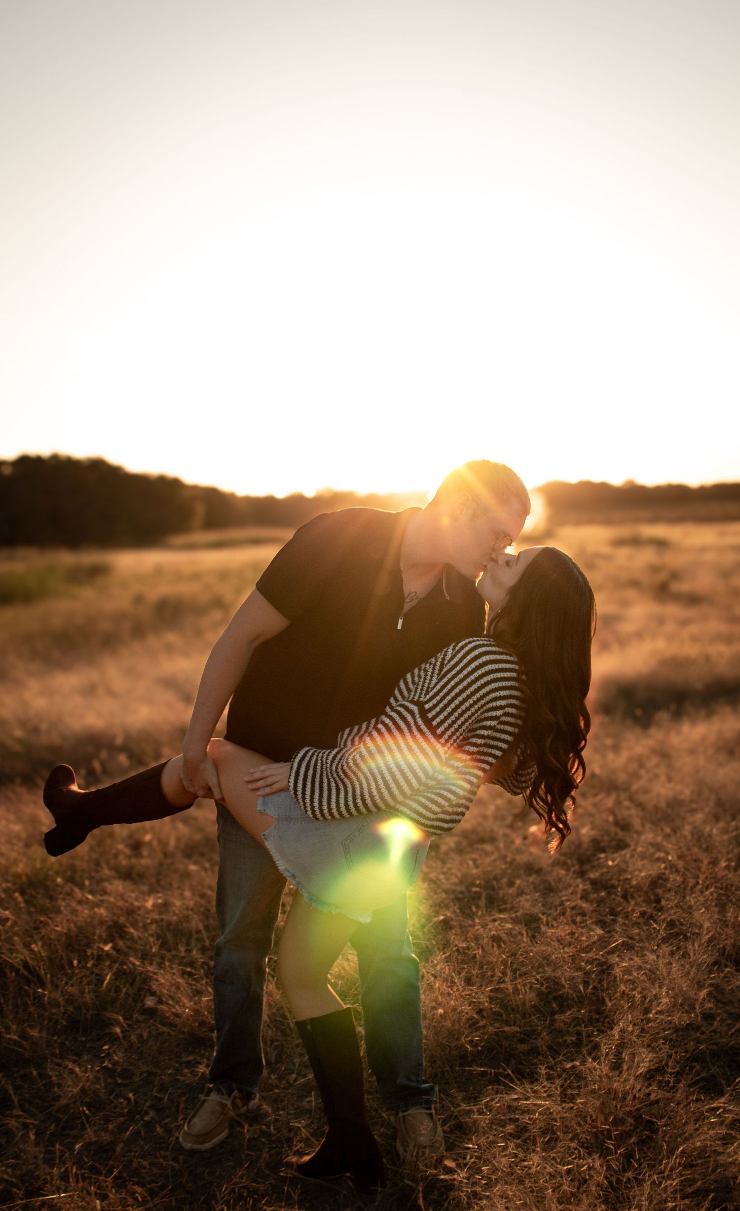 Couple sharing a romantic dip kiss in a golden field at sunset with warm lens flare lighting.