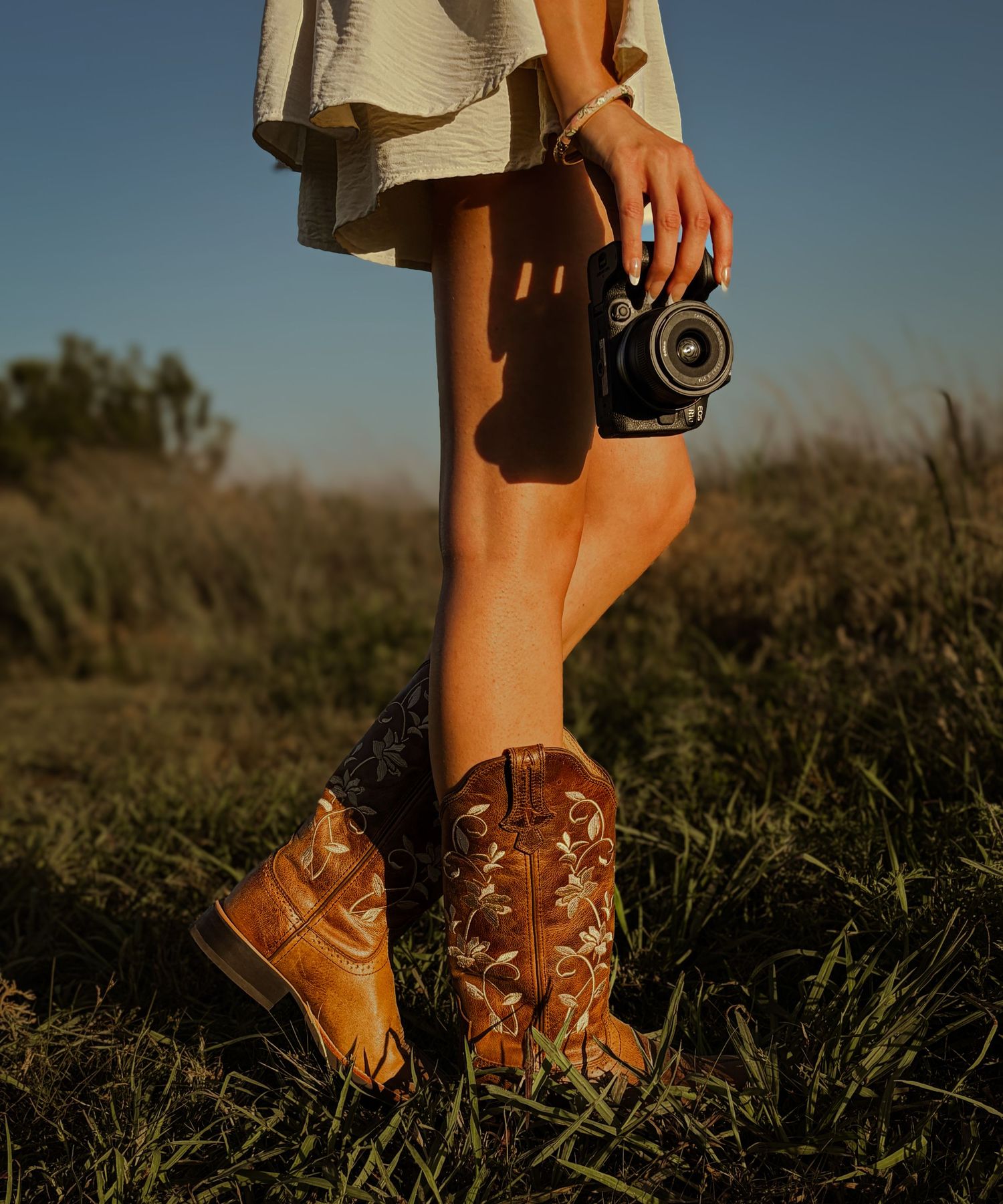 Woman in white dress and brown cowboy boots holding a vintage camera while standing in a grassy field.Branding inspo