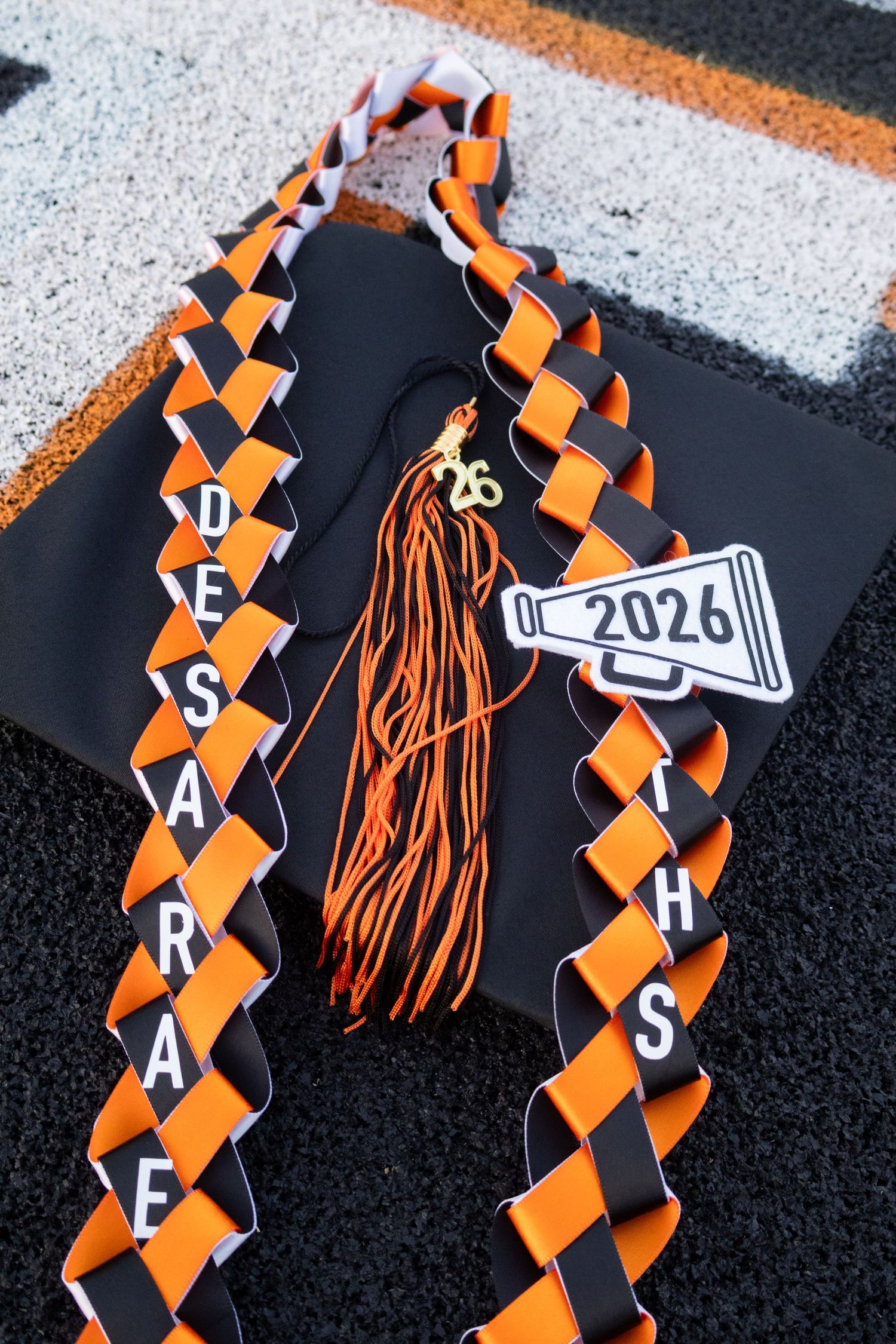 Black graduation cap with orange and black braided leis, tassel, and cheerleader megaphone decoration reading 2026. Senior Photos inspo