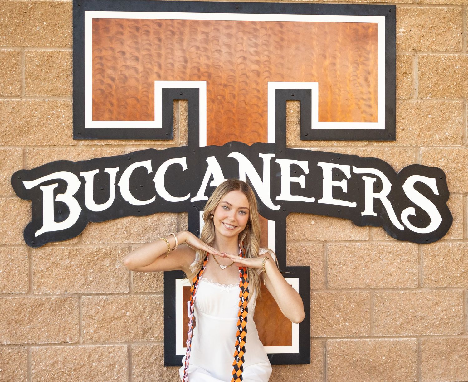 Young woman posing in front of a Buccaneers logo on a brick wall, making a hand gesture and smiling. Senior Photos Inspo
