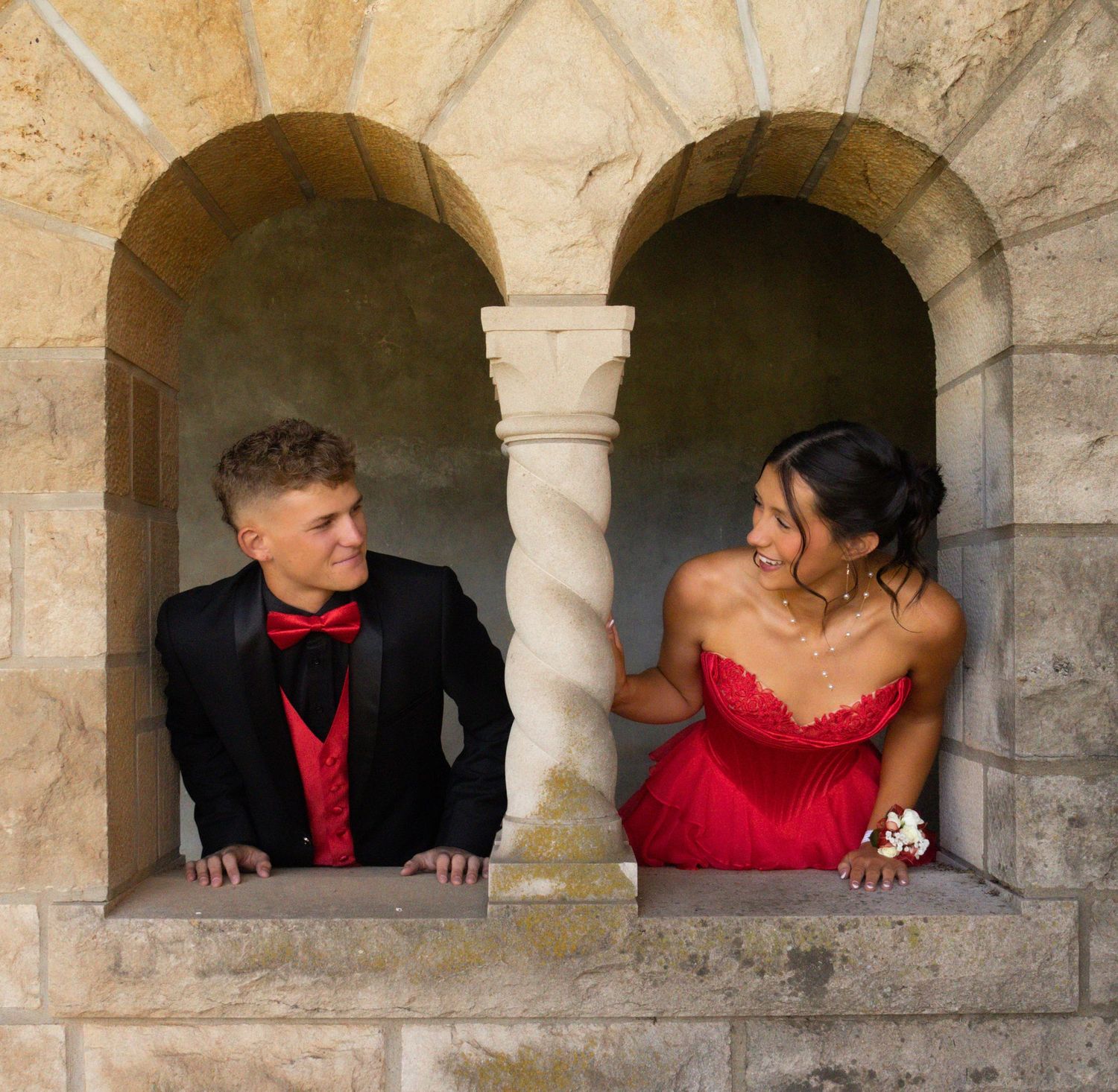 A couple in matching red and black formal attire pose in a stone archway, smiling at each other for prom photos.