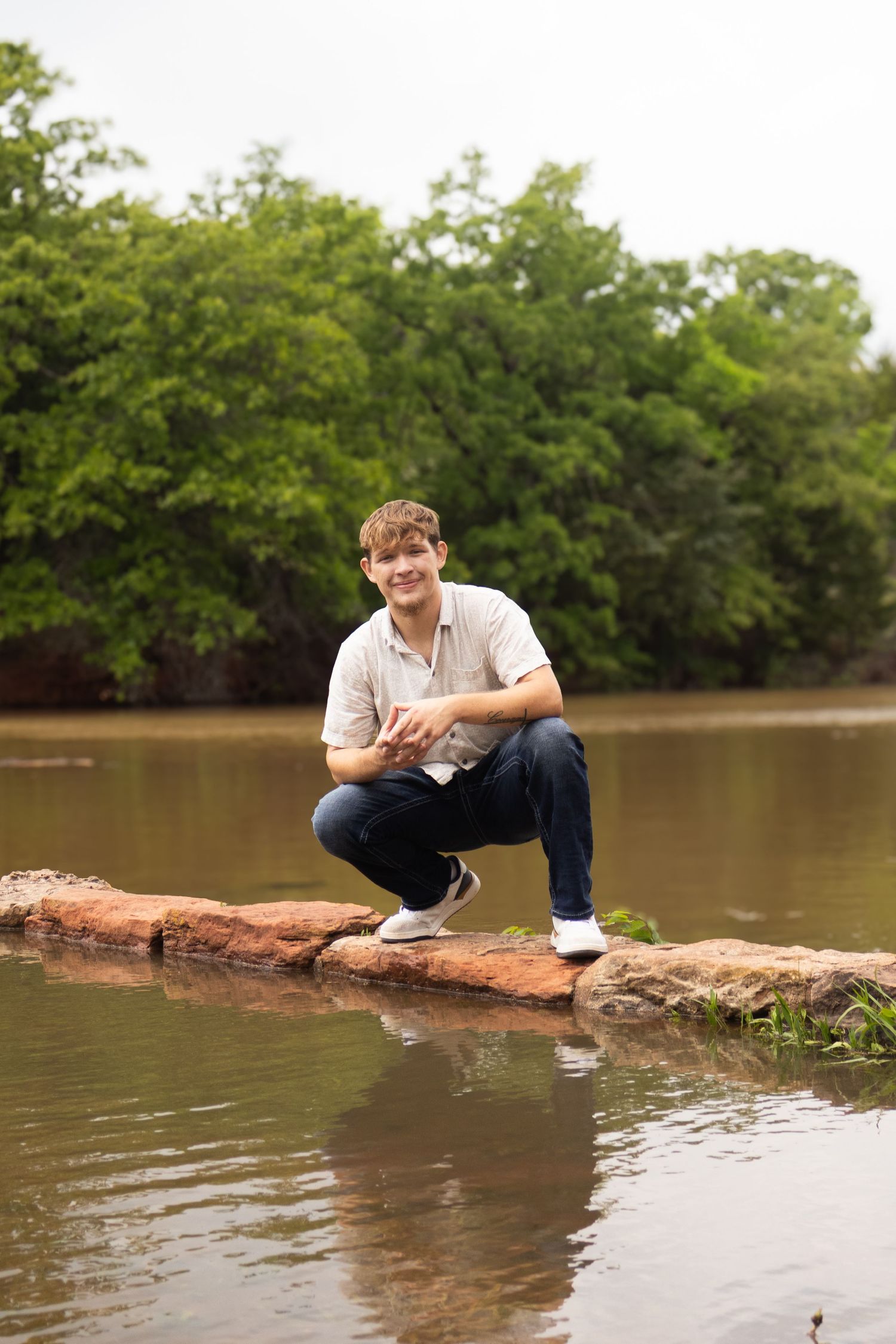 Young man crouching on rocks by a calm river, smiling, wearing jeans and a polo shirt, with lush green trees behind. Senior Photos inspo