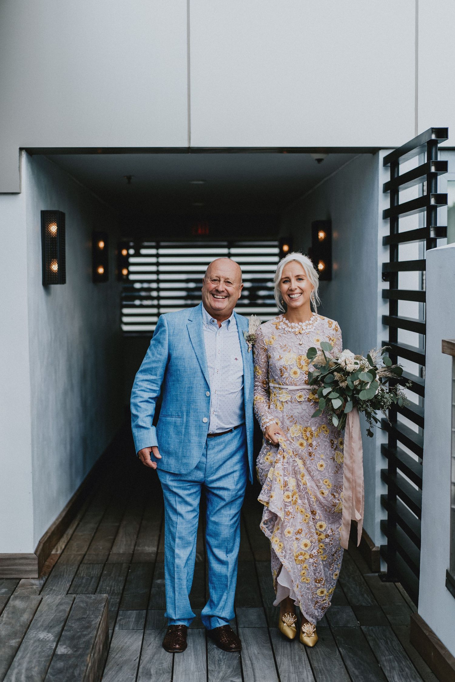 Smiling couple in wedding attire, man in blue suit and woman in floral gown holding bouquet, standing in venue doorway.