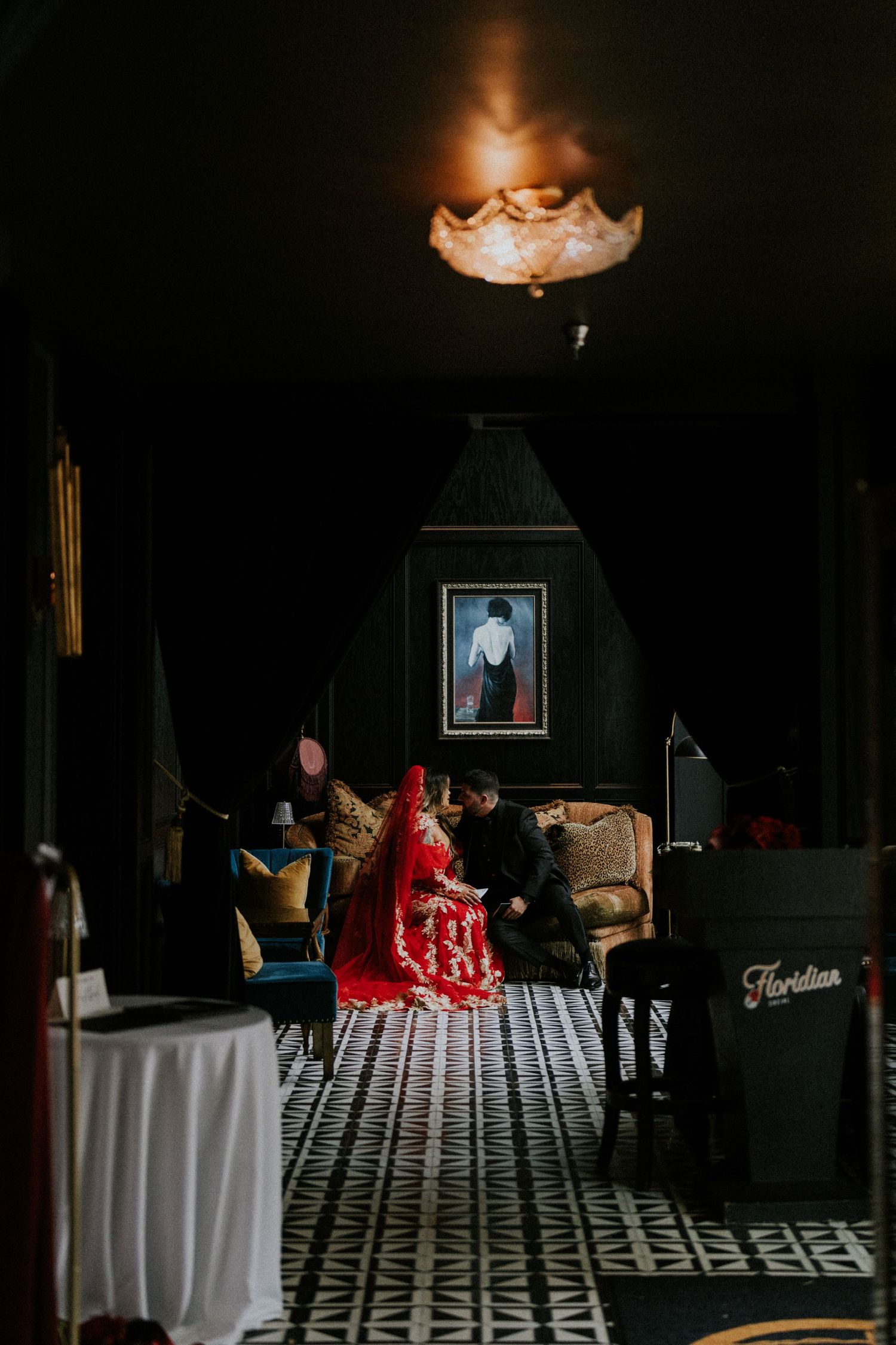 Bride in red Chinese wedding dress and veil seated with groom in dark moody venue interior.