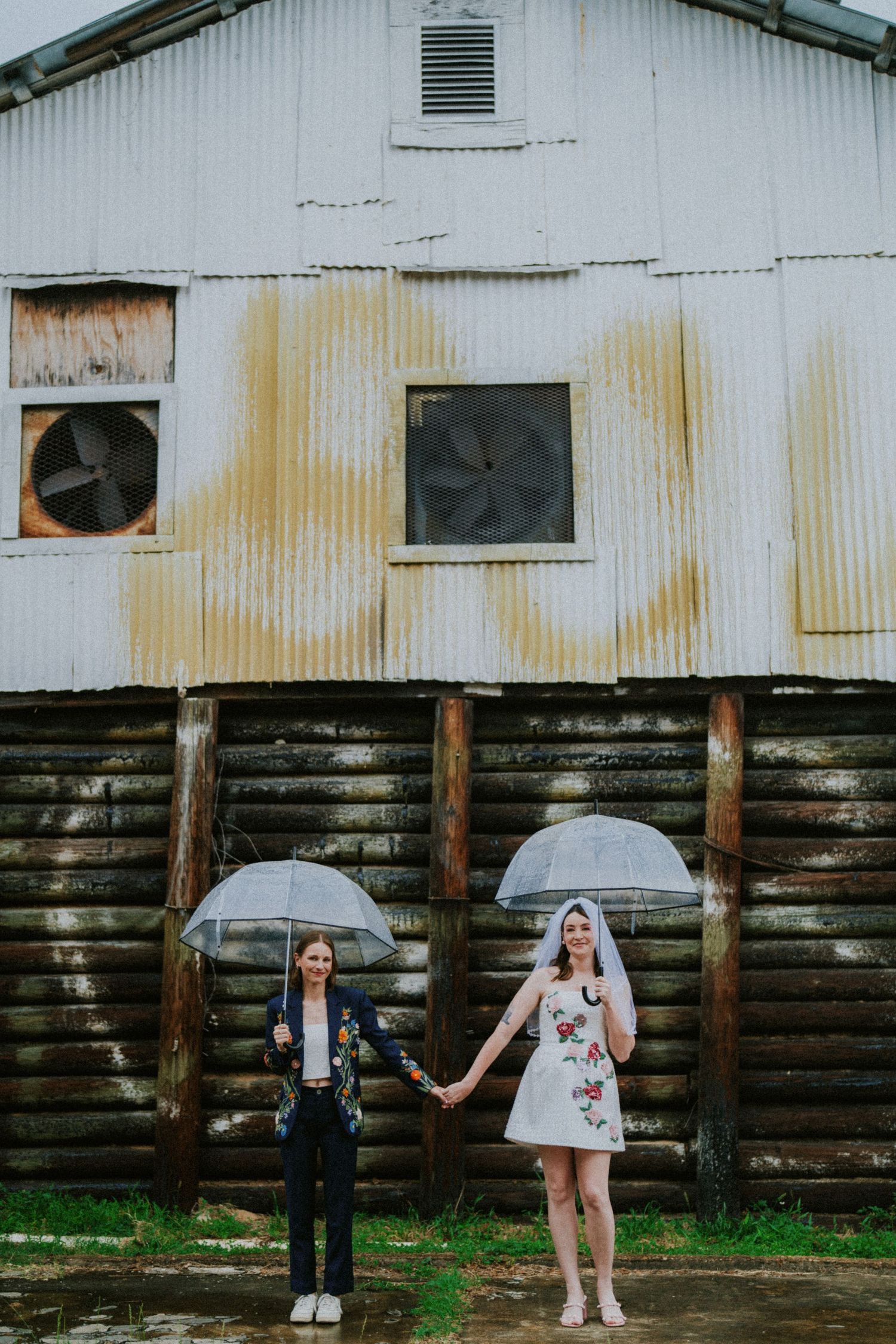 Two brides holding clear umbrellas in the rain, standing hand in hand before a rustic corrugated metal barn.
