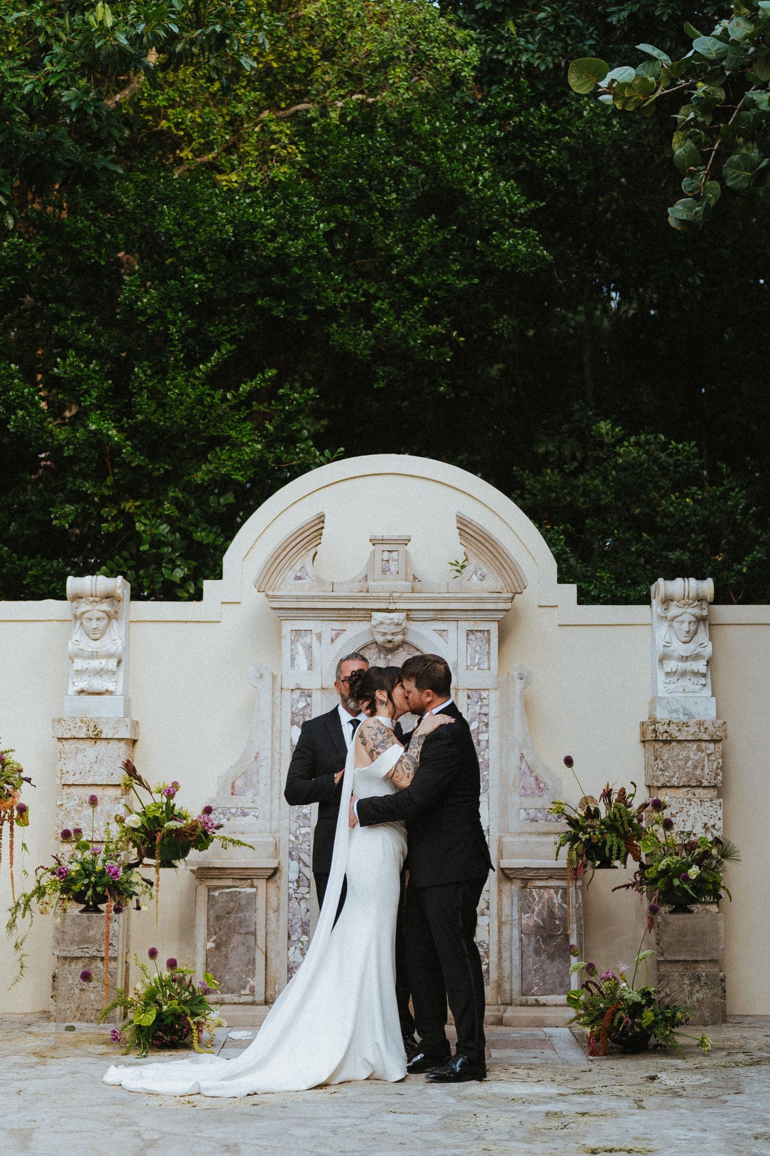 Couple shares a kiss at the altar in front of an ornate white stone arch adorned with floral arrangements.
