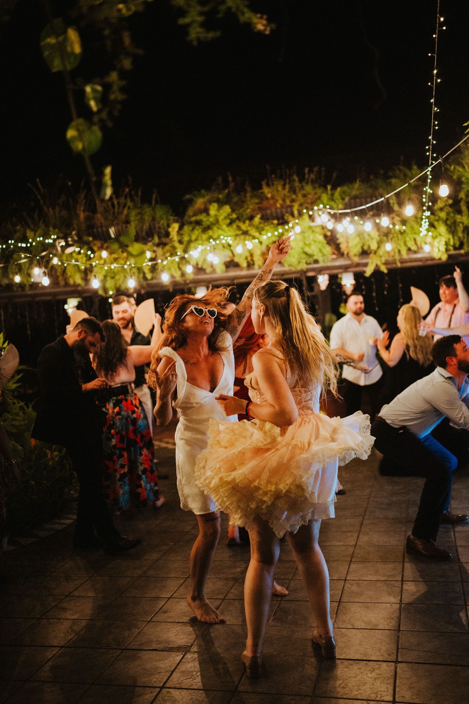 Two women in white dresses dance joyfully at an outdoor evening party with string lights and guests in background.