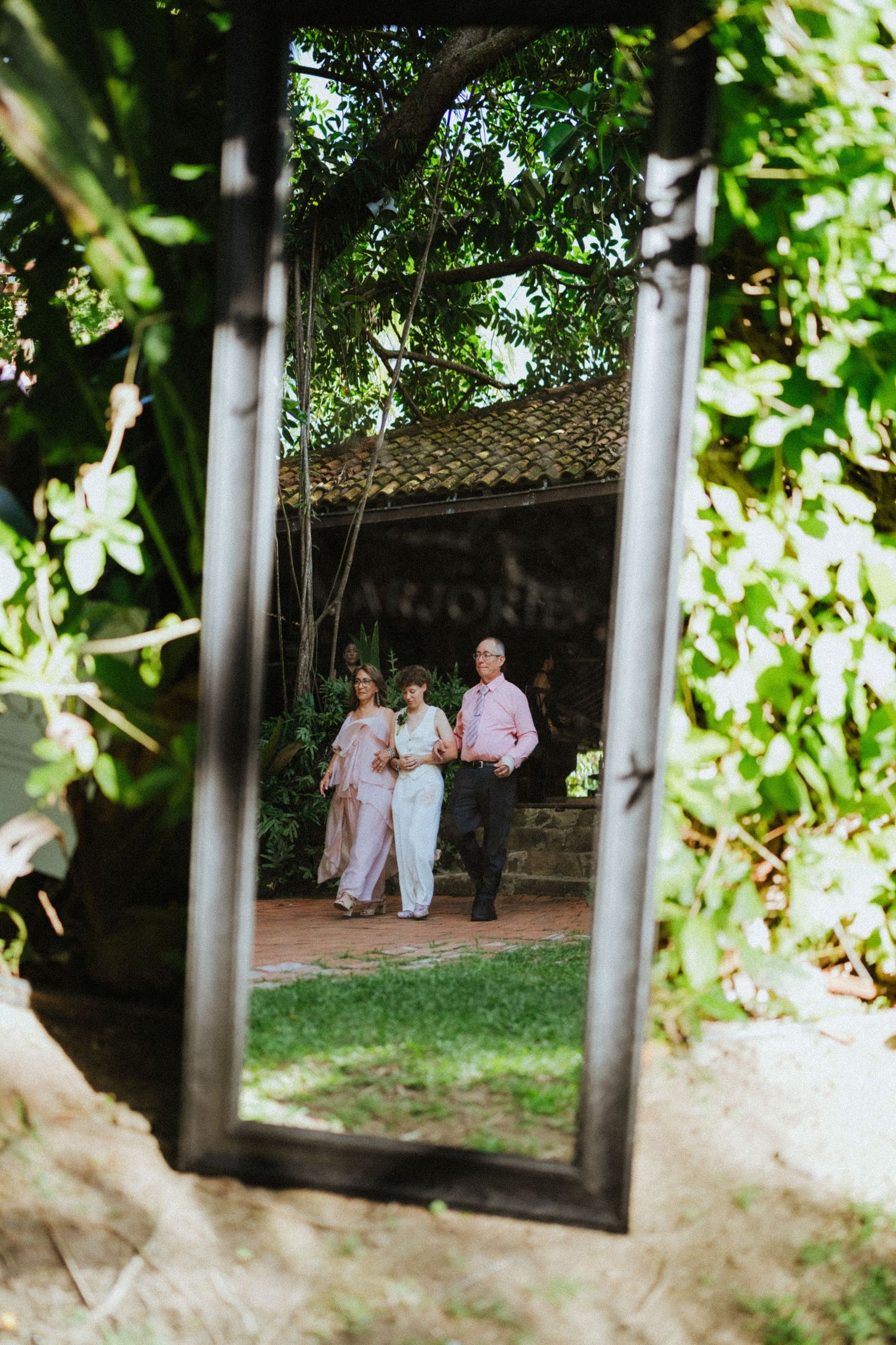 Mirror reflection of a bride and her parents walking through a lush garden venue on wedding day.