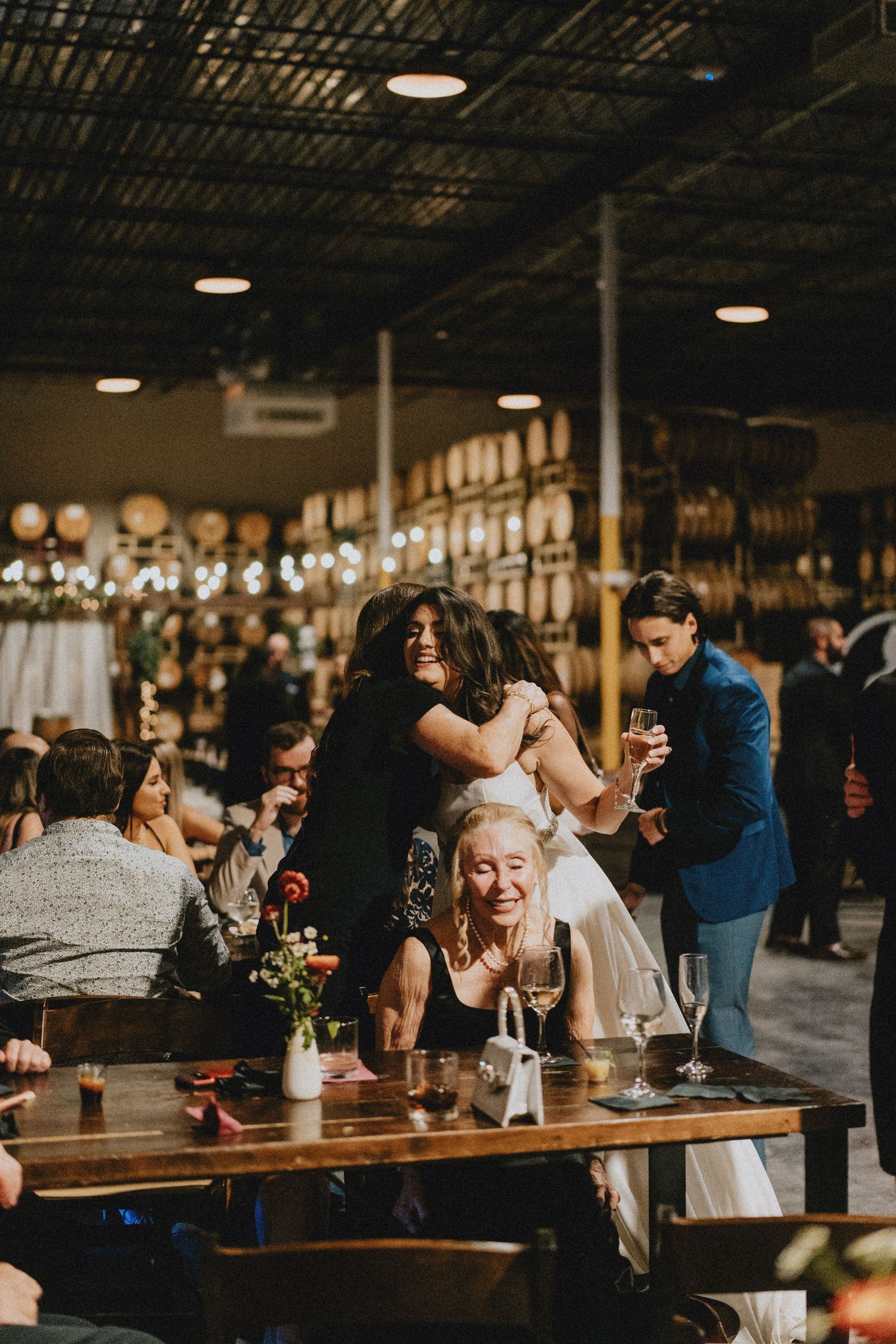 Wedding guests laugh and embrace warmly at rustic distillery reception with barrel-lined walls and string lights.