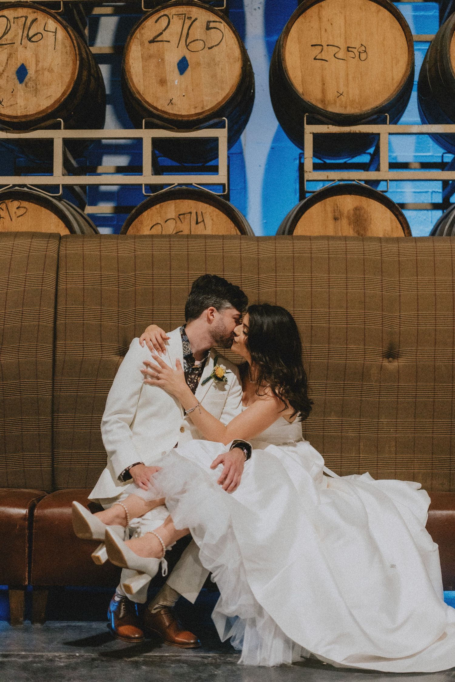 Newlyweds share a passionate kiss while sitting on a leather couch surrounded by numbered wooden barrels.