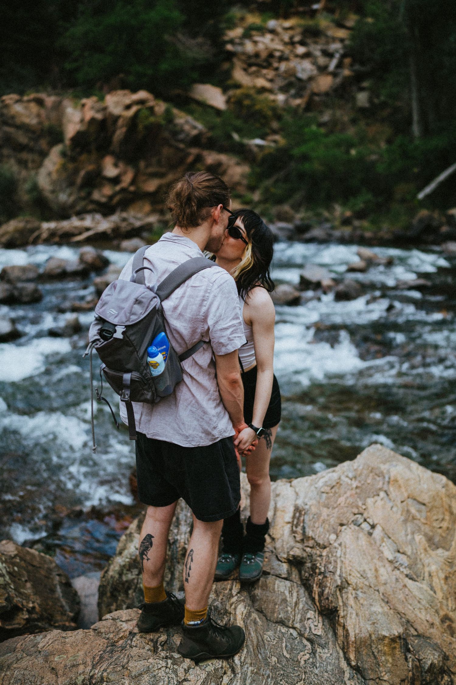Couple kissing on rocky riverbank with rushing mountain stream and boulders in background.