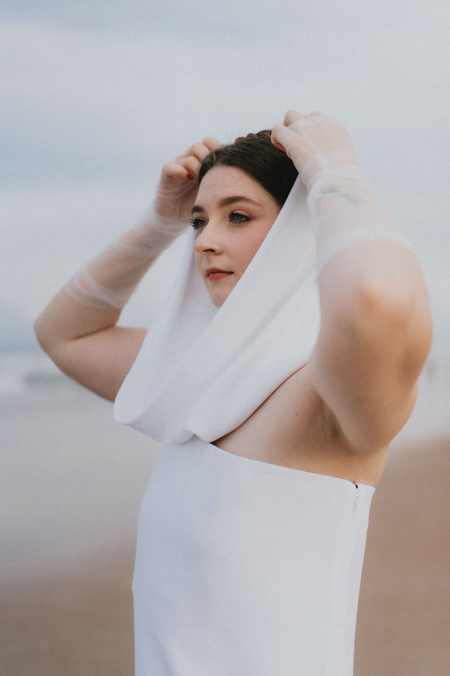 Bride on a beach adjusts her white veil over her head while wearing a sleeveless white wedding gown.