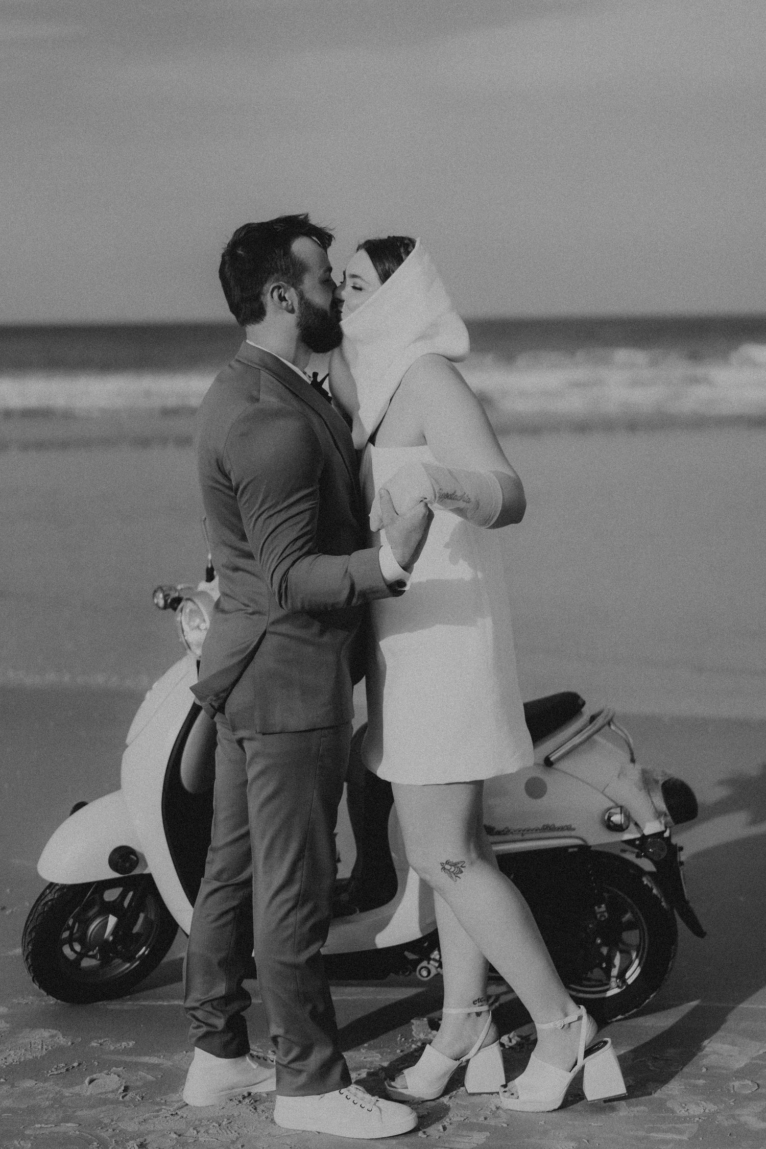 Black and white photo of a couple kissing on a beach beside a vintage Vespa scooter.