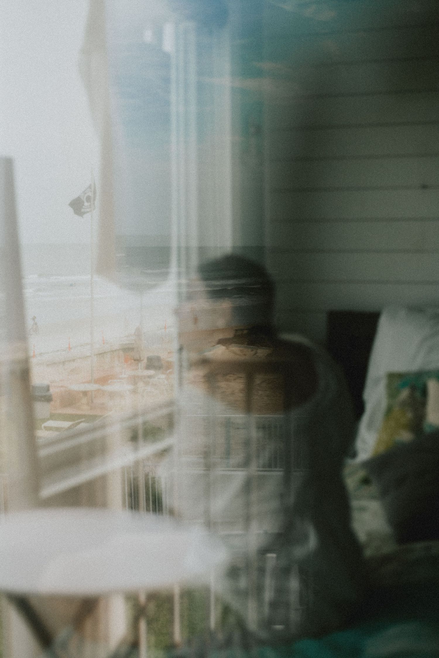 Blurry reflection of a person in a window overlooking a beachside scene with white siding and ocean views.