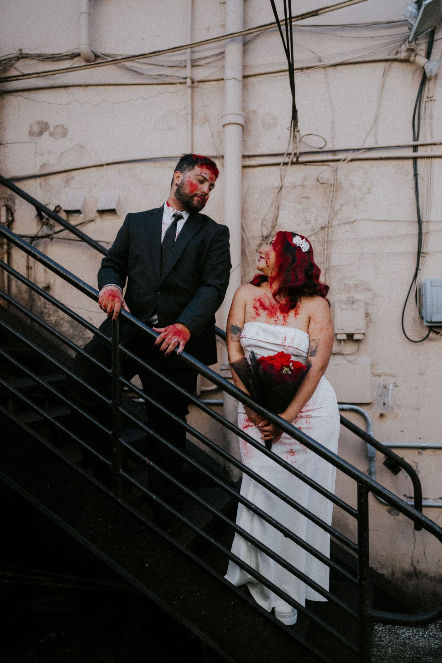 Couple in zombie wedding costumes with fake blood on stairs, groom in black suit, bride in white dress.