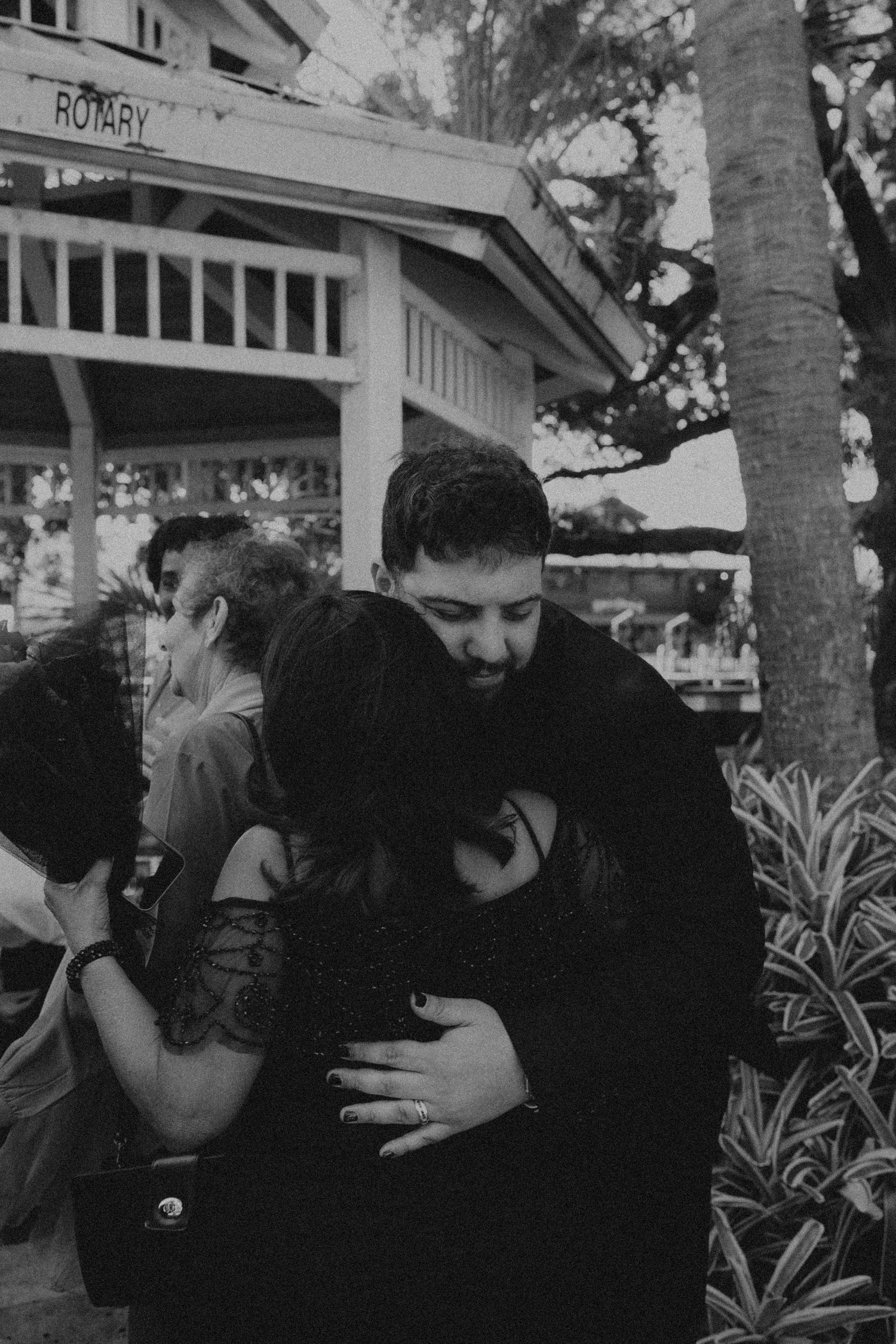 Black and white photo of a bearded man tenderly embracing a woman outside a Rotary pavilion surrounded by trees.