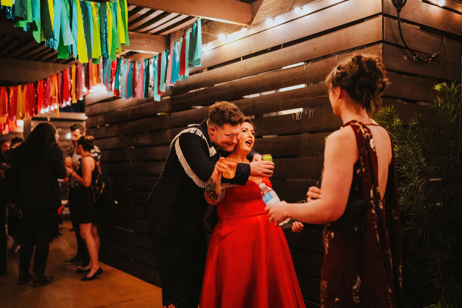 Guests in formal attire socializing at a festive indoor party venue with colorful hanging decorations.