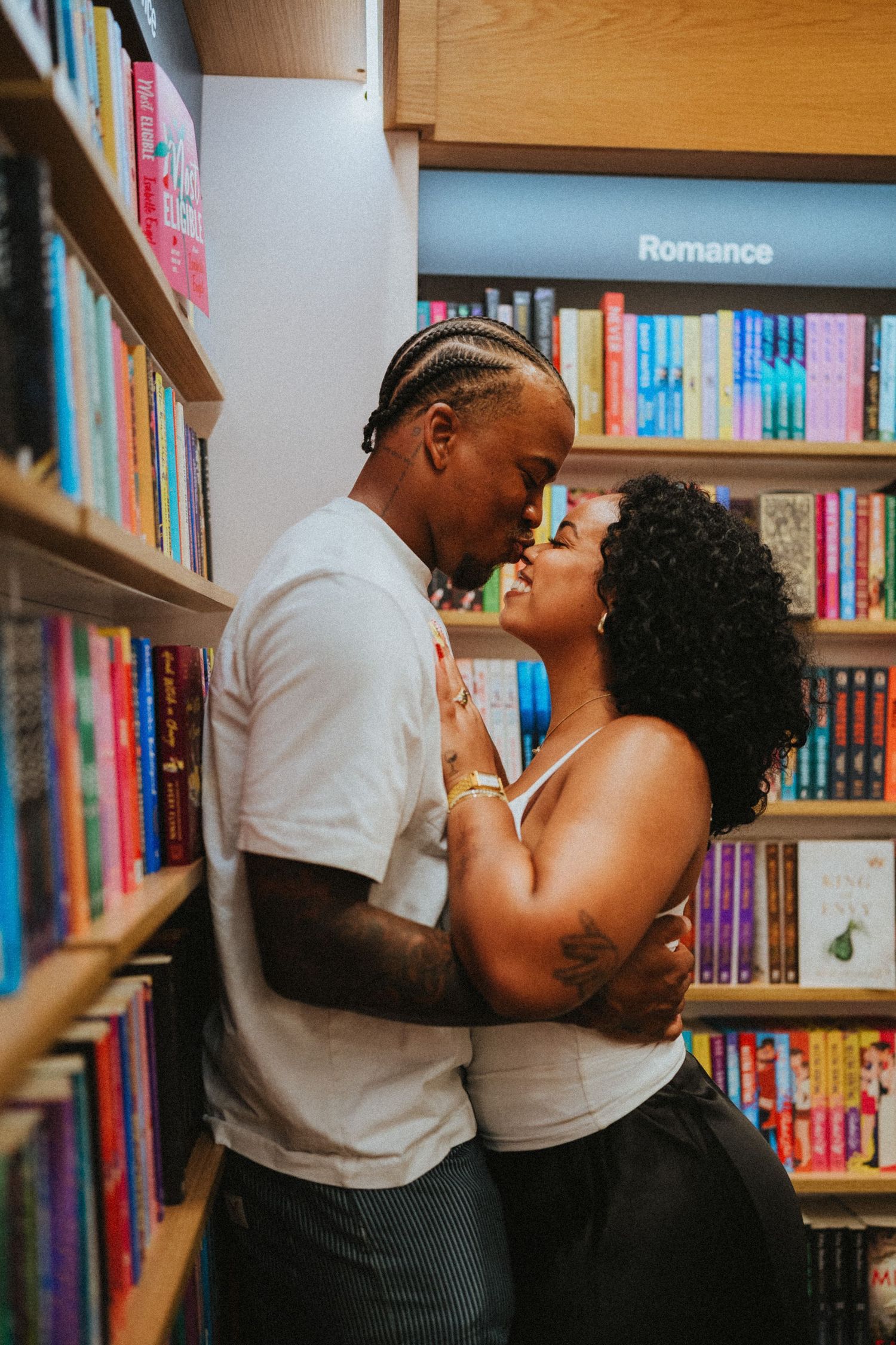 A couple shares a romantic kiss in a bookstore beneath a Romance section sign, surrounded by colorful shelves.