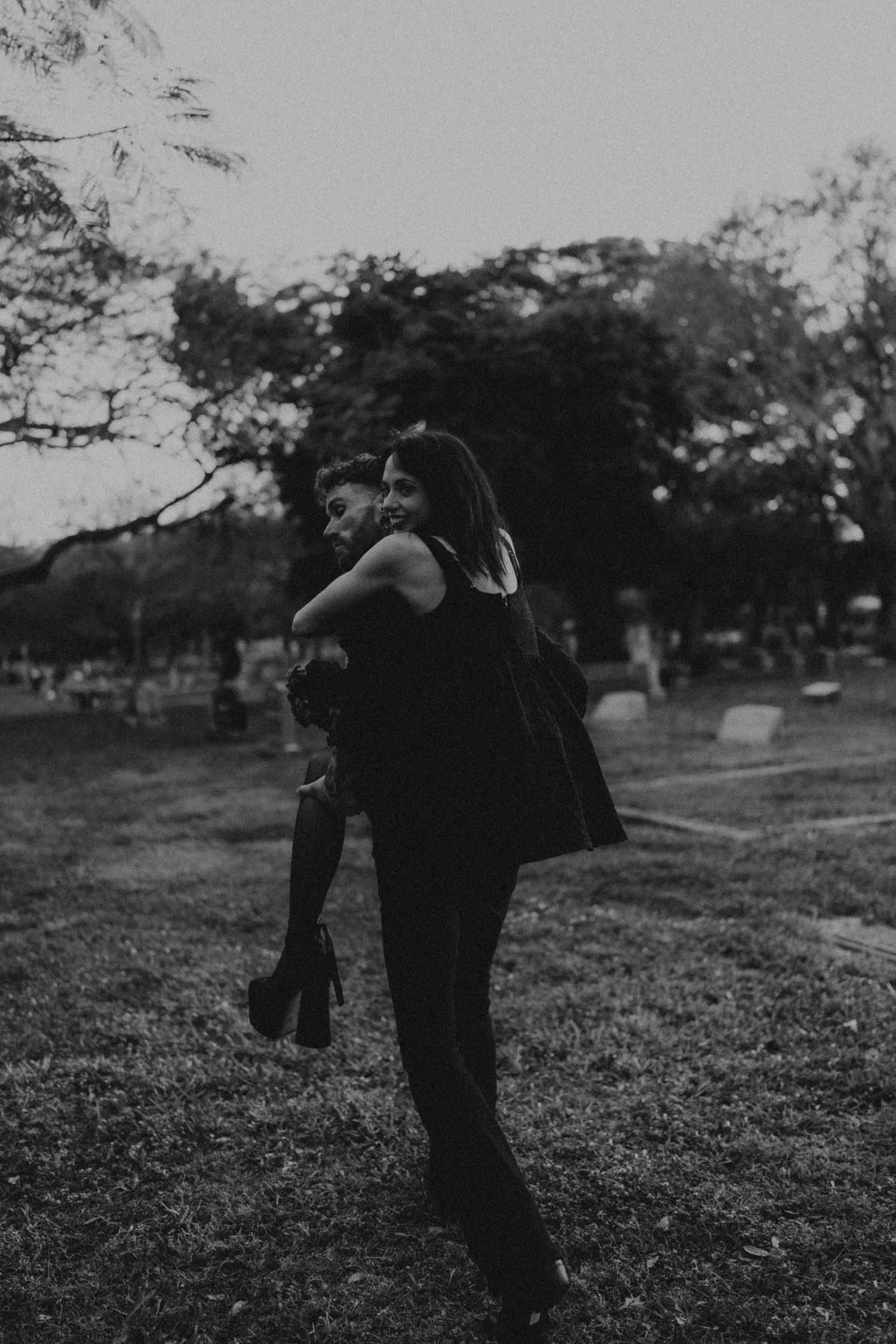 Black and white photo of a couple sharing a piggyback ride in a park at dusk, surrounded by trees.