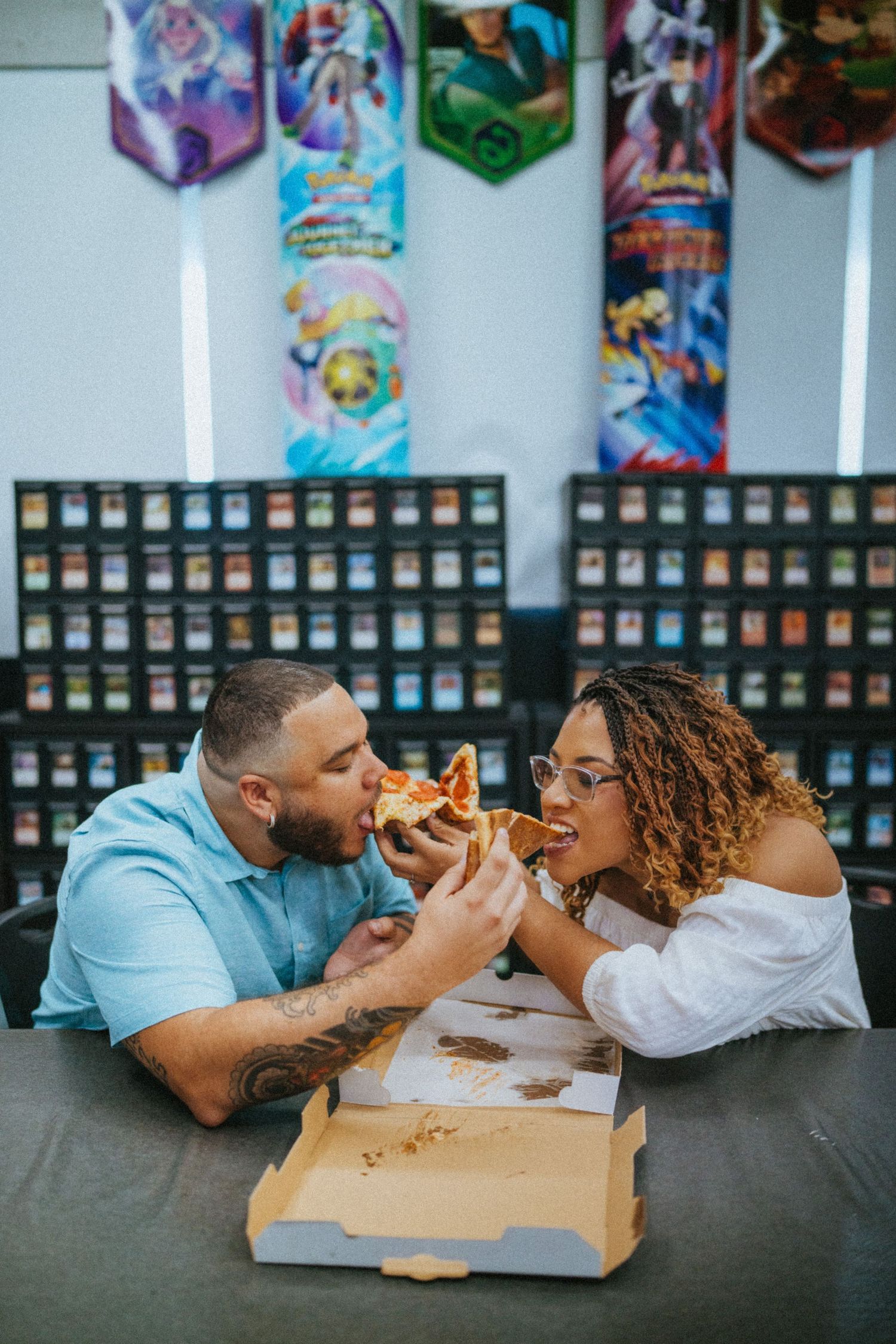 Couple sharing a pizza slice at a gaming card shop with Pokemon banners and card displays in the background.