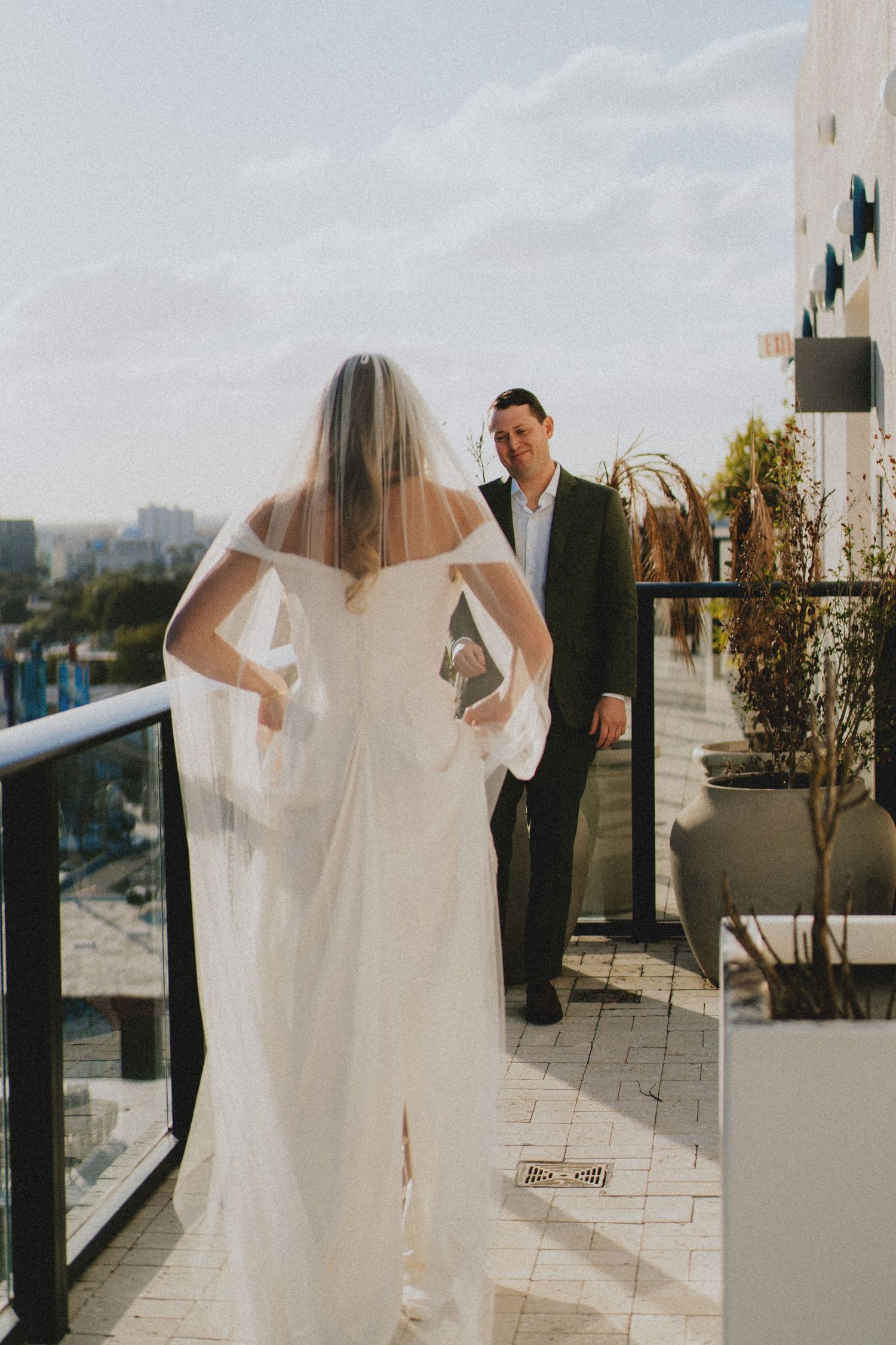 Bride in white gown and veil walks toward groom on rooftop balcony during wedding first look.