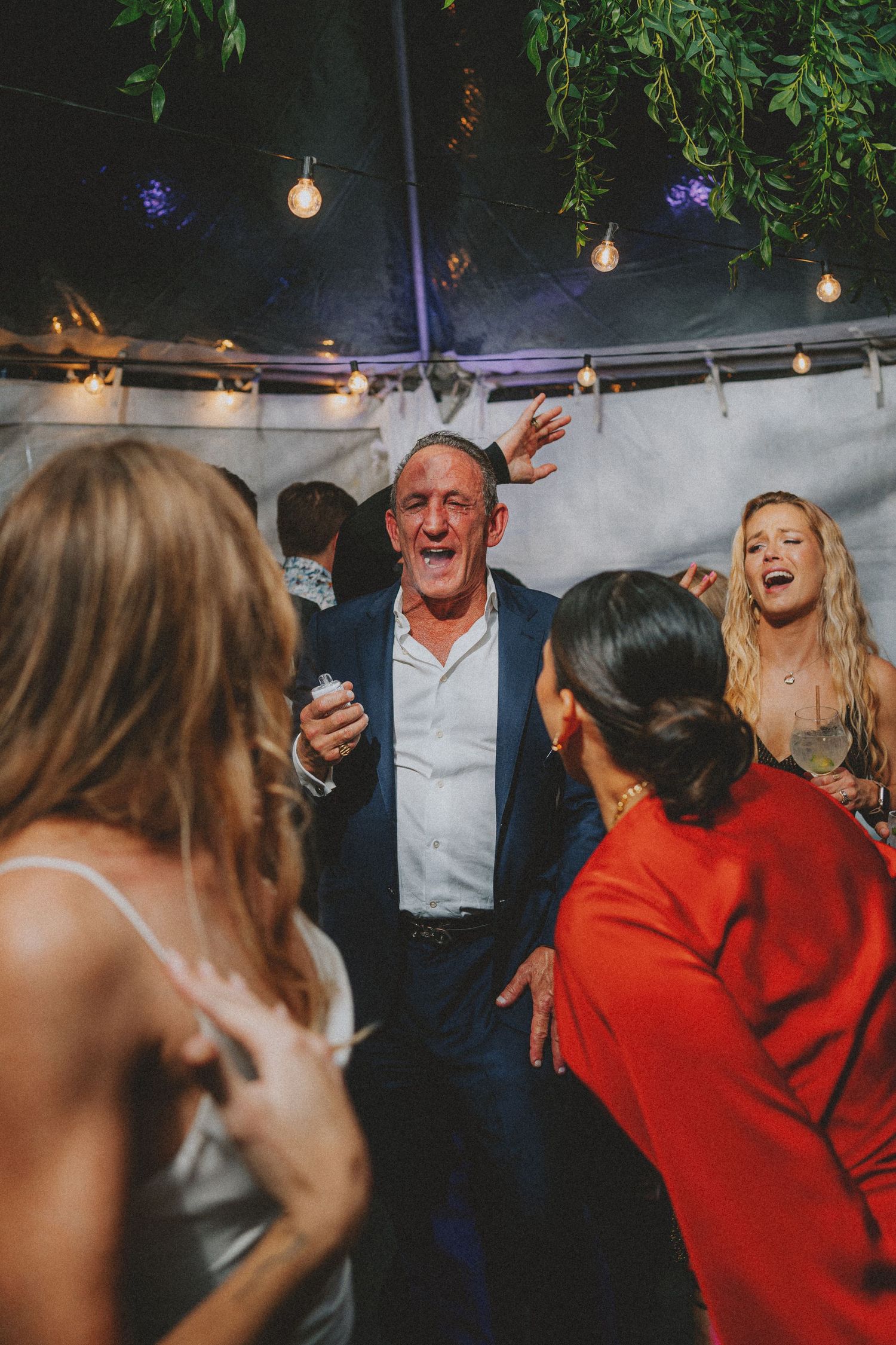 Older man dancing energetically with wedding guests under string lights at reception.
