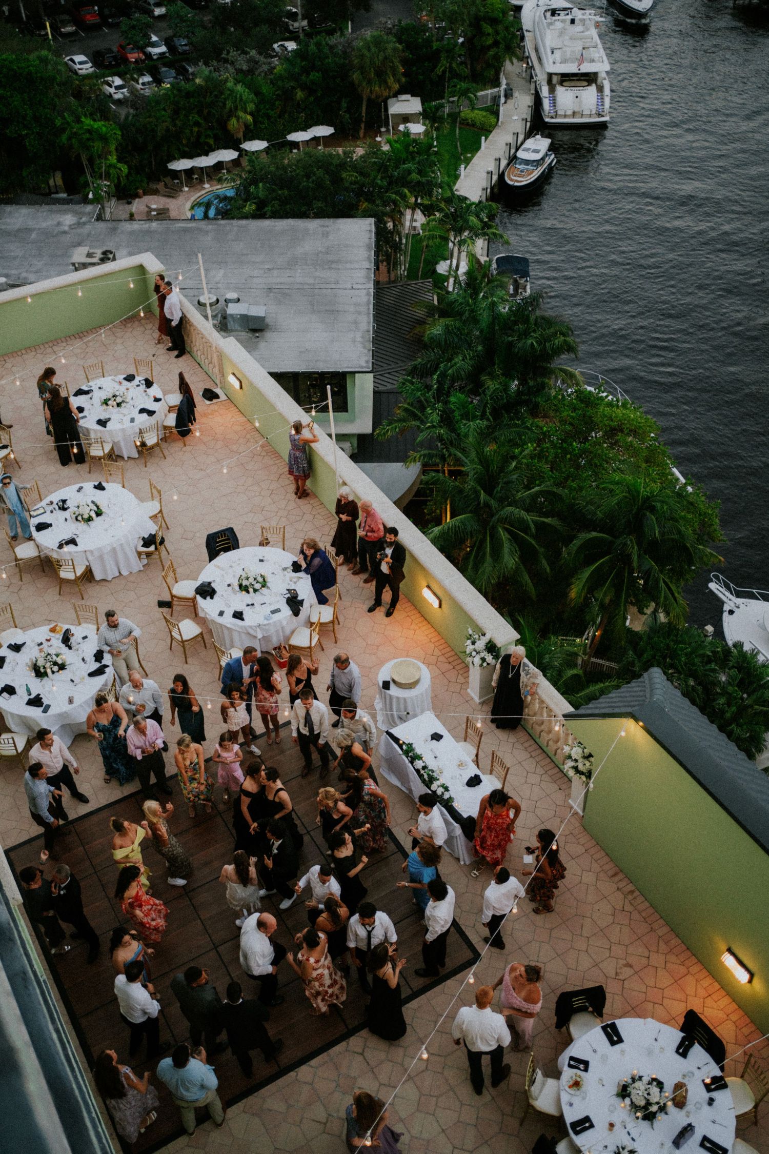 Aerial view of an outdoor waterfront reception venue with round tables and guests mingling on a terrace near boats.