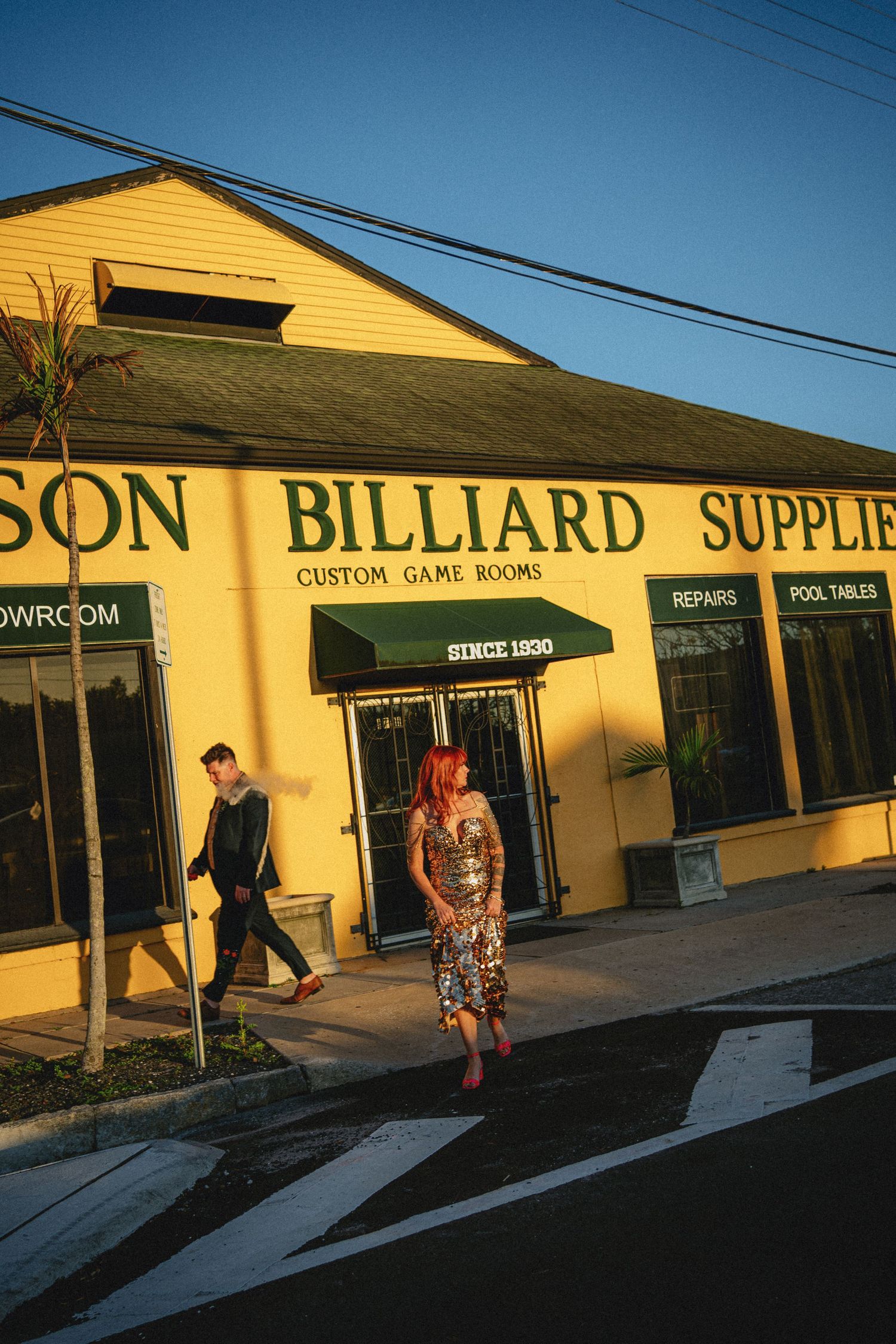 Couple poses outside a yellow billiard supplies shop with green awnings at golden hour.