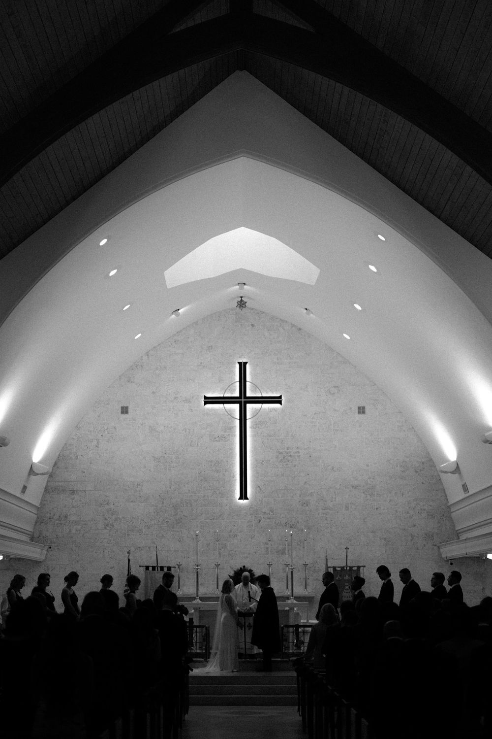 Black and white photo of a church ceremony with a large illuminated cross on the altar wall.