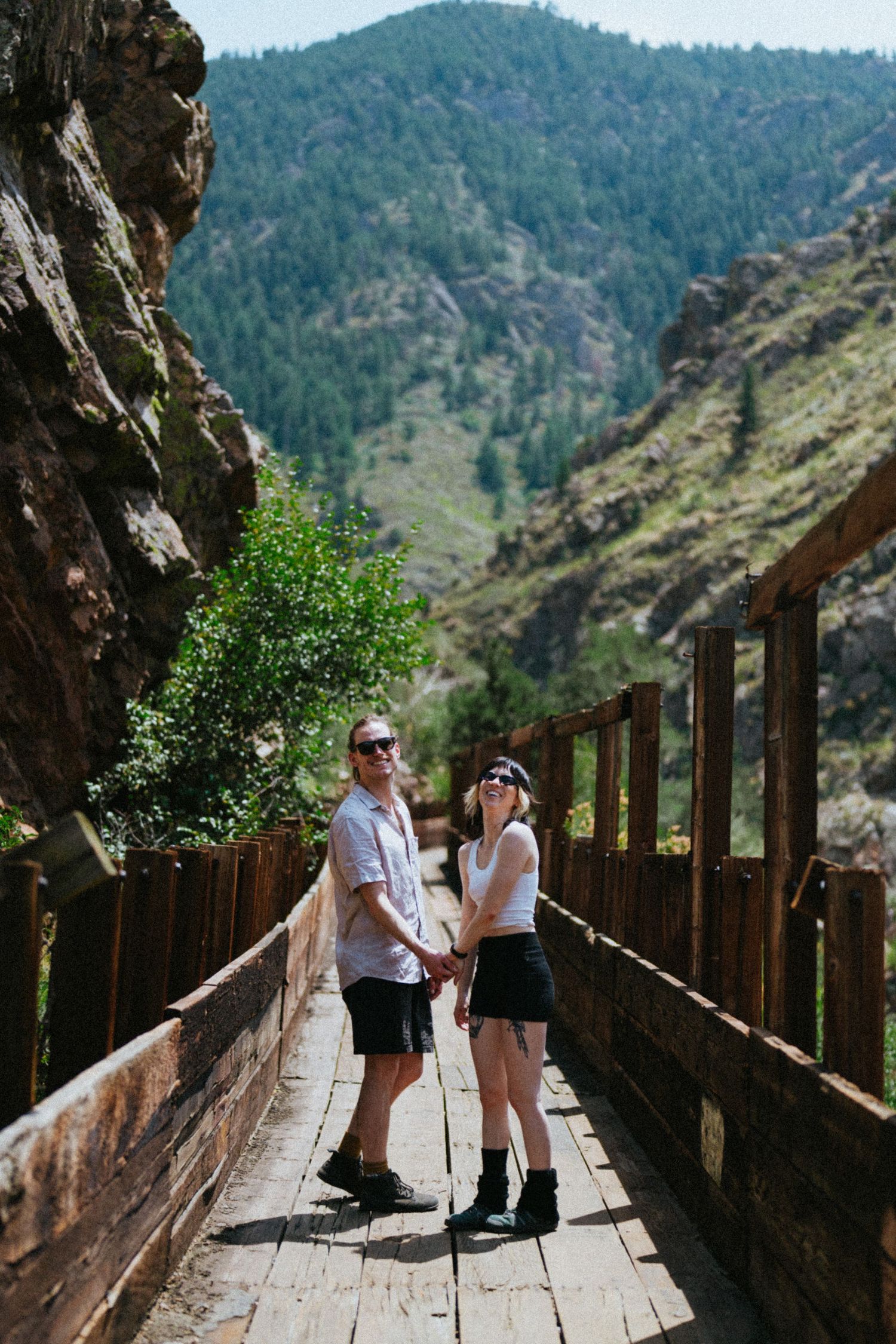 Couple holding hands on a wooden bridge walkway with mountain forest backdrop on a sunny day.