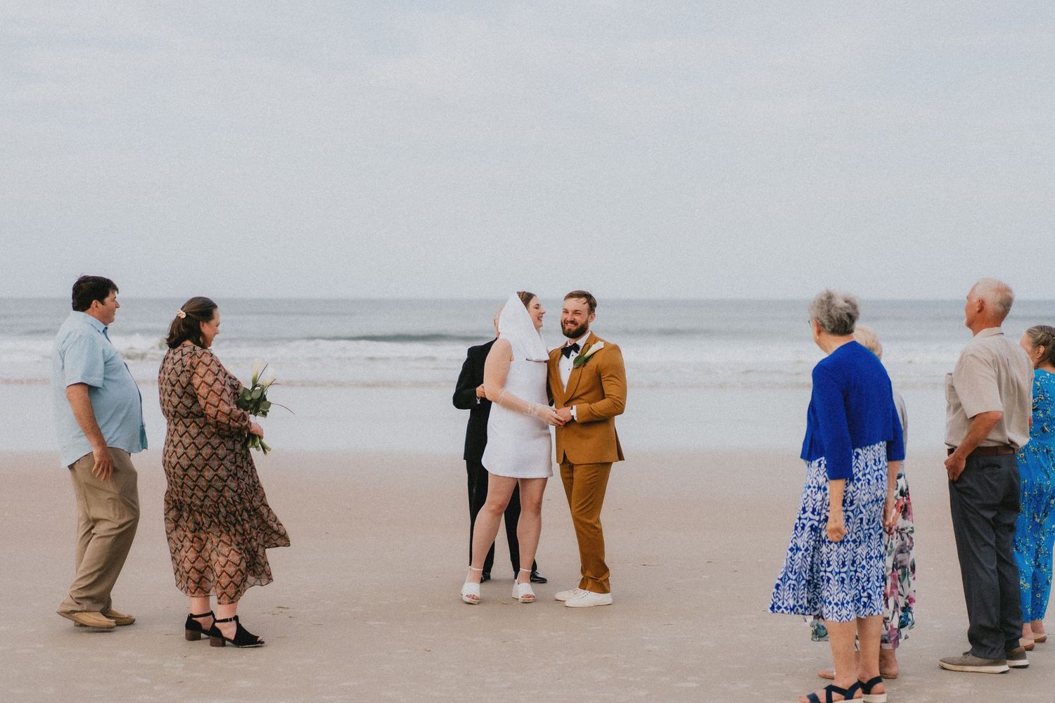 Intimate beach wedding ceremony with bride and groom exchanging vows surrounded by small group of guests on sandy shore.