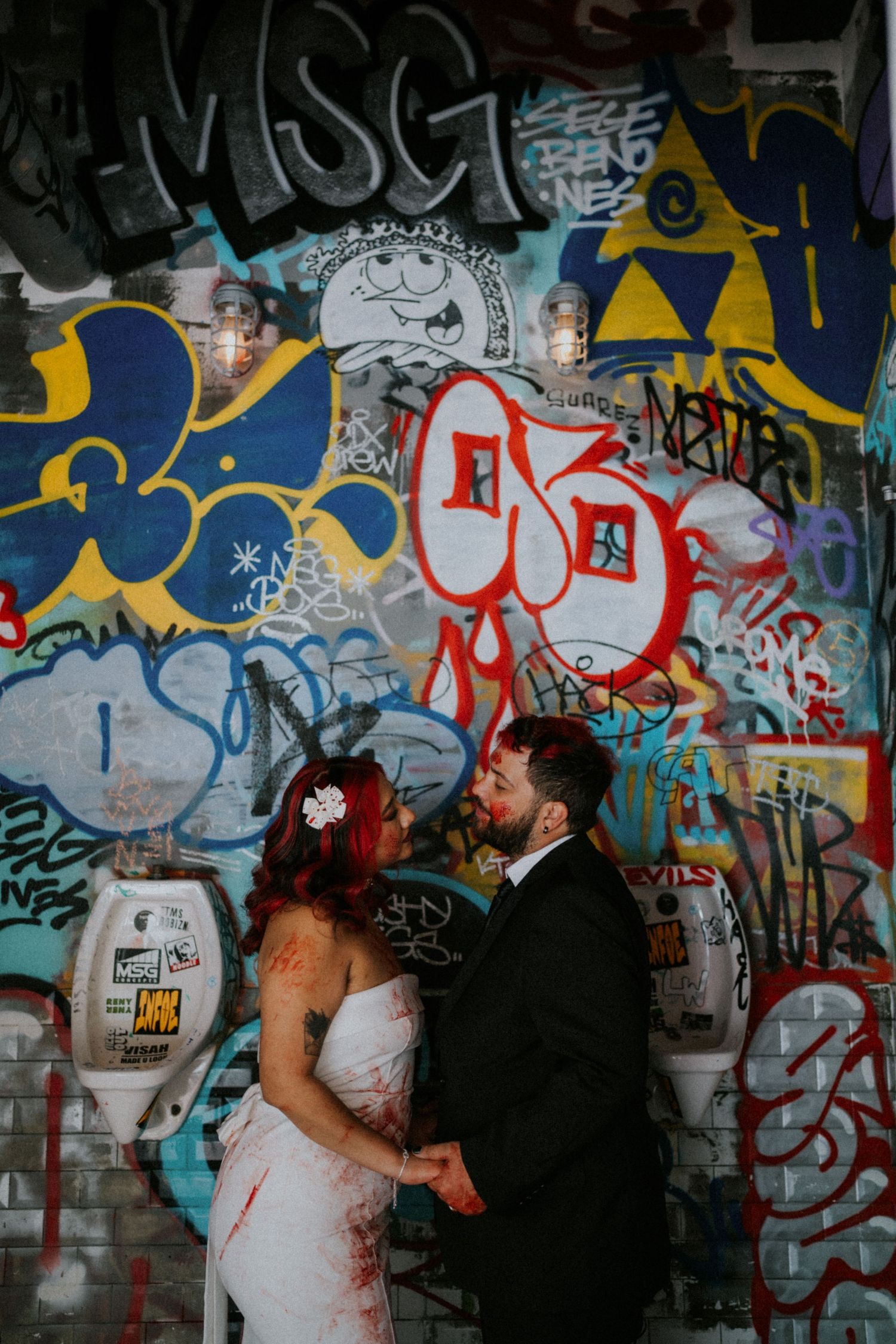 Wedding couple embraces in front of colorful graffiti wall, bride in red-stained white dress.