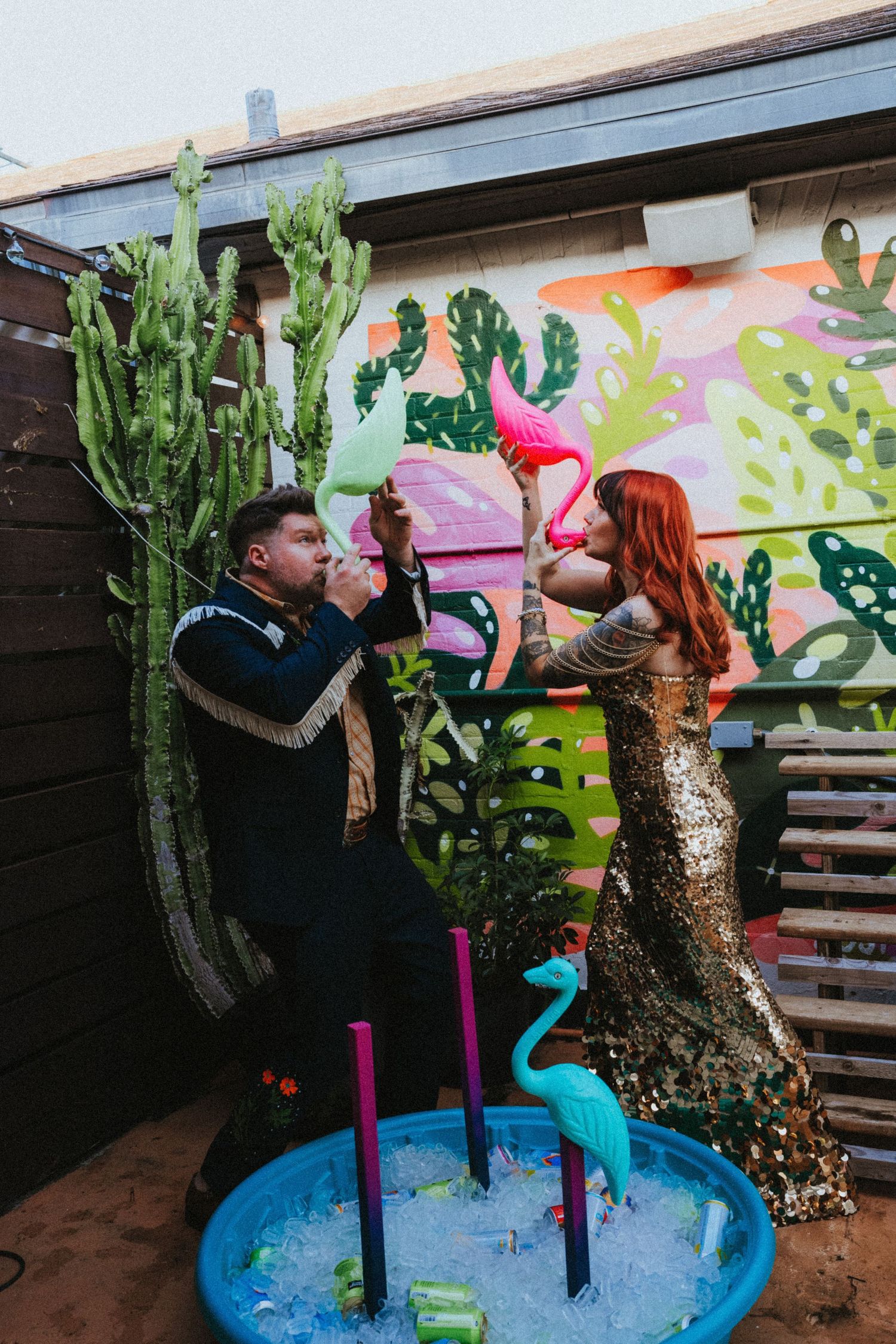 Couple in festive attire play with inflatable flamingo near a colorful cactus mural and kiddie pool.