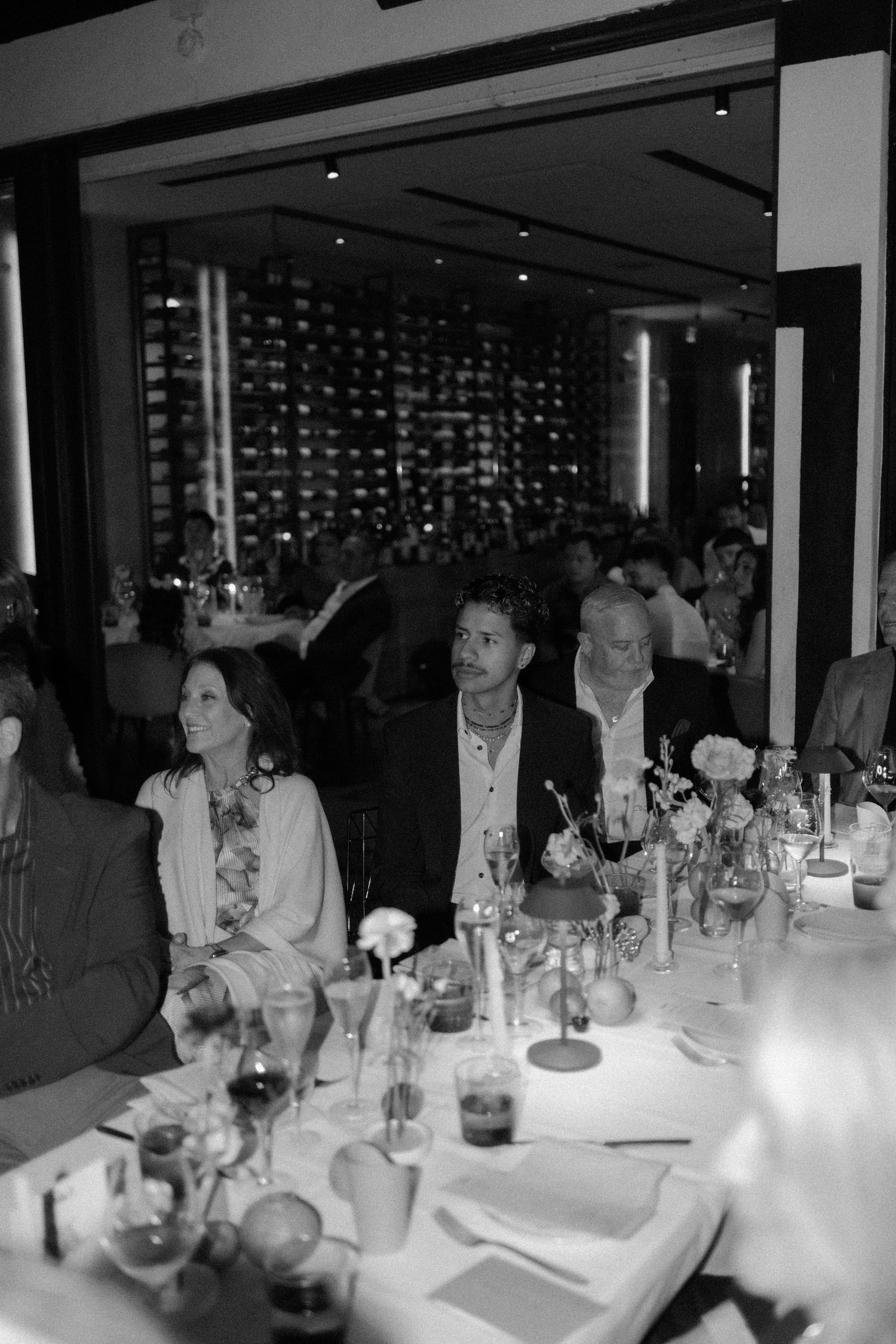 Wedding guests seated at elegantly set dinner tables in a dimly lit restaurant, black and white photo.