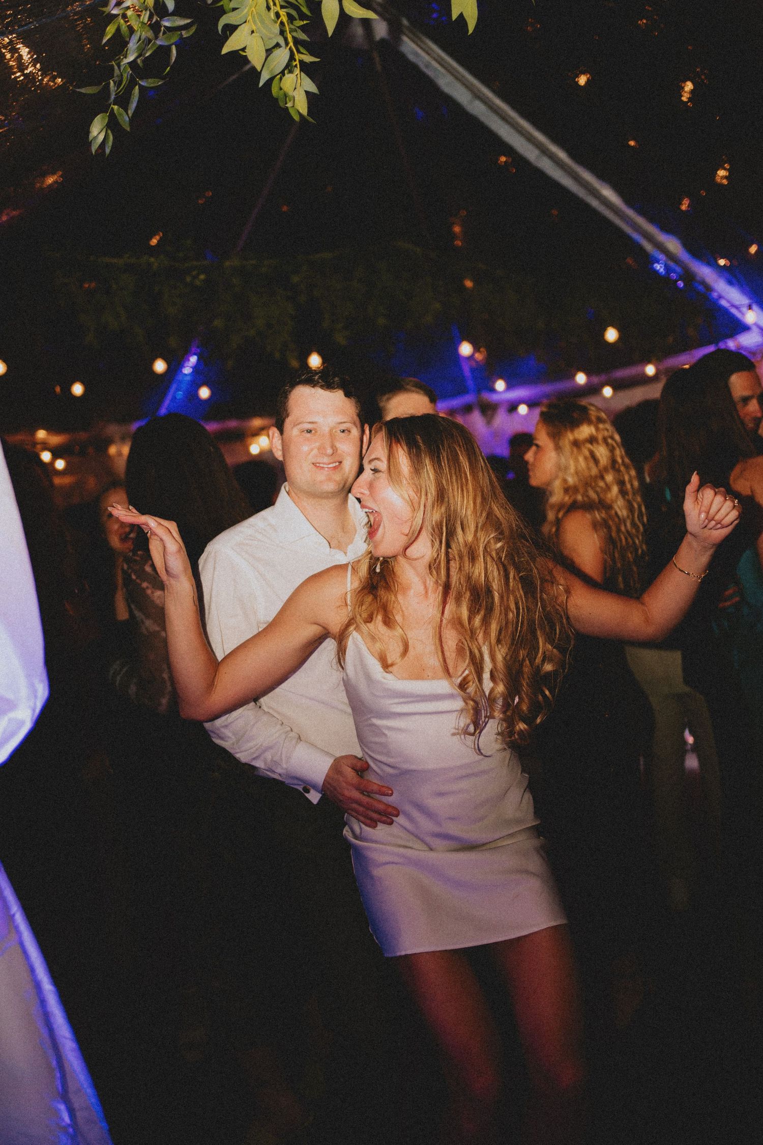 Bride in white mini dress dances closely with partner at a festive outdoor wedding reception.