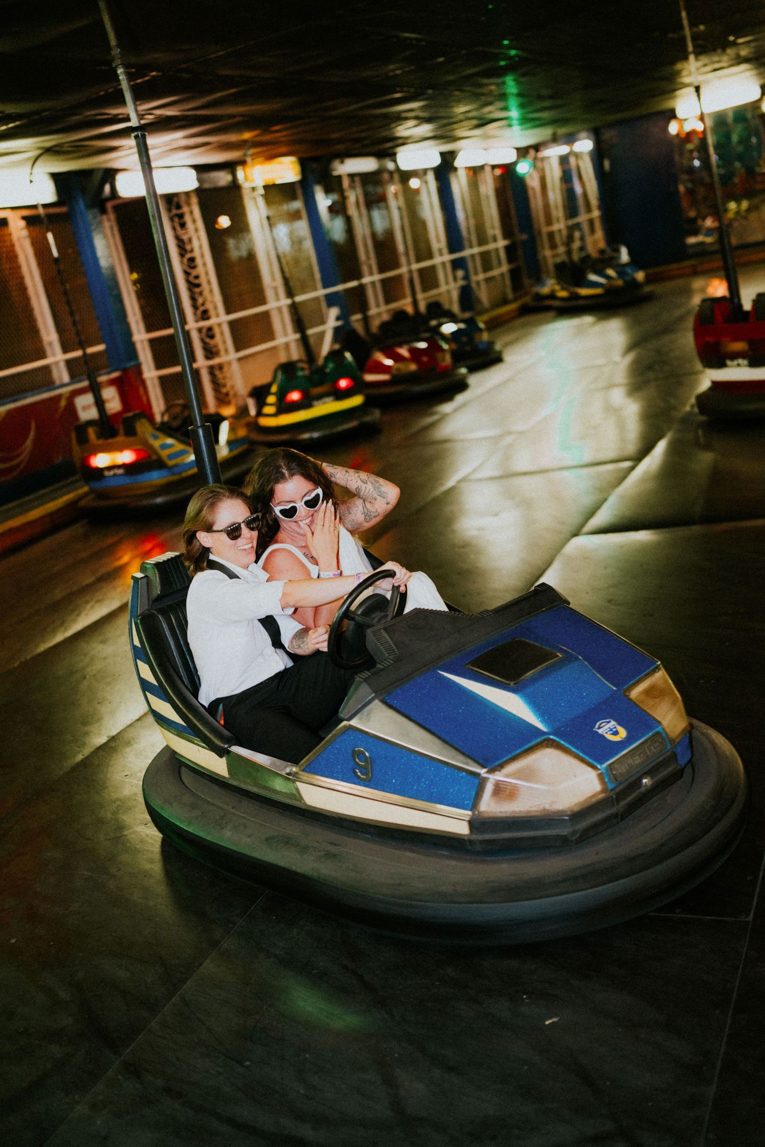 Two young women wearing sunglasses laugh while riding together in a blue bumper car at an amusement park.