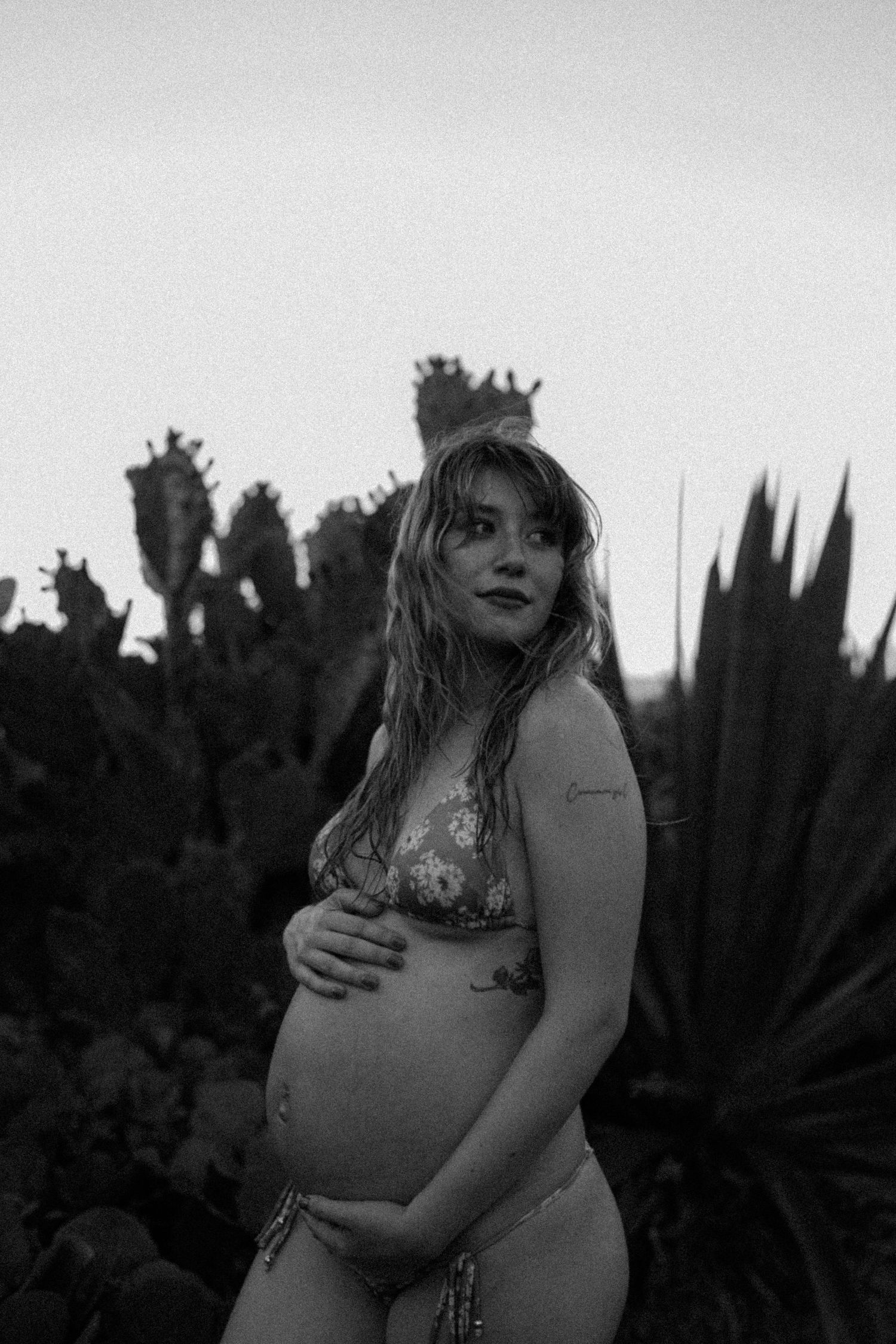 Black and white photo of a pregnant woman in a bikini cradling her baby bump outdoors among cacti at dusk.